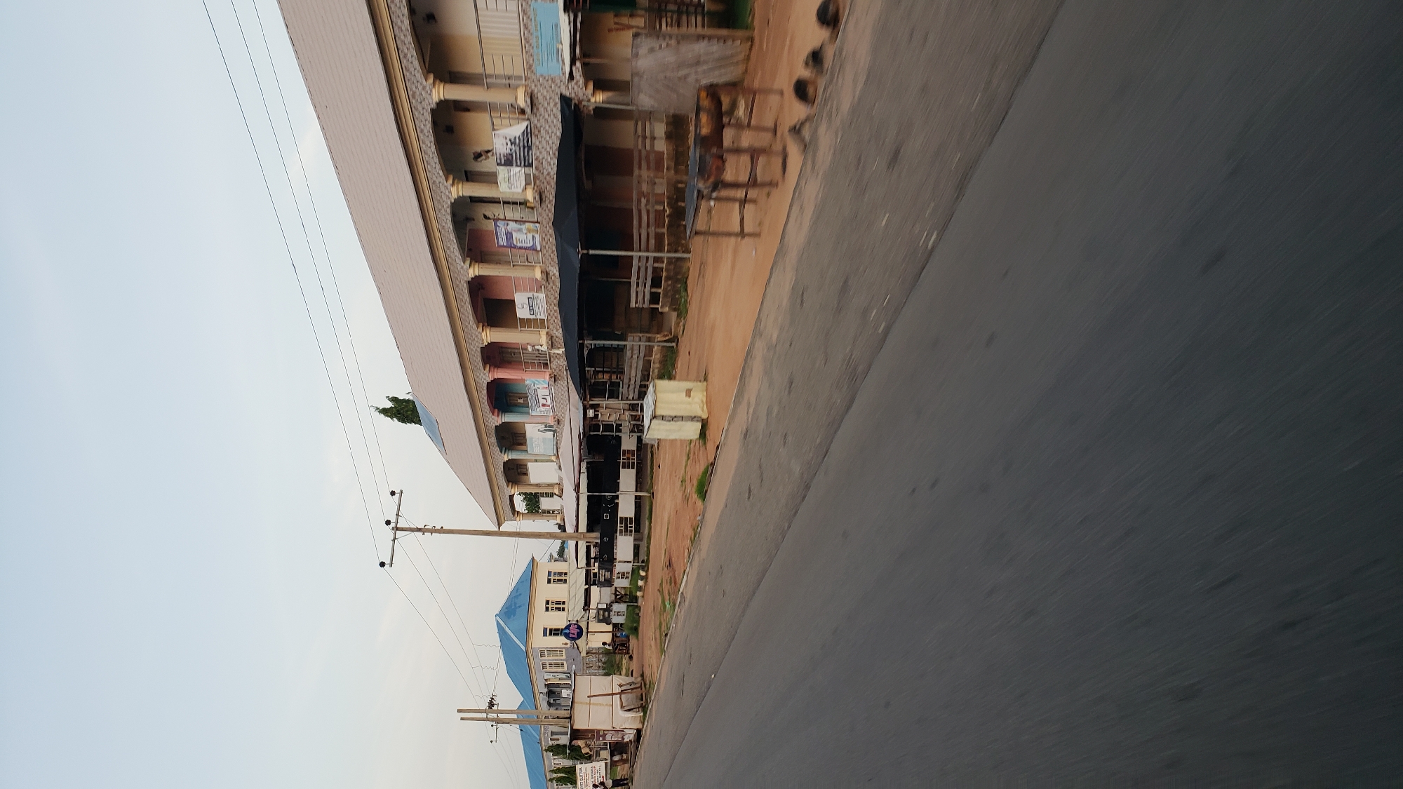The image depicts a street scene in Jalingo, Nigeria, with a two-story building prominently featured on the right side of the frame. This building has a light-colored, tiled roof and a facade adorned with columns painted in pastel shades of pink, blue, and yellow. The upper level of the building features a balcony with railings, and several windows or doorways are visible. Signs are posted on the upper facade, some of which appear to be advertisements or informational notices.
In front of this building, a covered area with a dark fabric roof is set up, supported by wooden posts and railings. Further to the left, another building with a blue, pitched roof is visible, also with multiple stories and windows. A utility pole with electrical wires stretches across the scene.
The foreground is dominated by a paved road, blurred due to motion, suggesting the image was taken from a moving vehicle. The side of the road is dirt, with some scattered debris and what appear to be wooden structures, possibly stalls or seating areas. One such structure in the middle distance consists of a metal grill over a fire pit, placed on a wooden table. A small, light-colored rectangular object, possibly a shed or container, is situated on the dirt beside the road. A few people are visible in the distance, near the buildings on the left. The sky above is clear and light, indicating it is daytime.