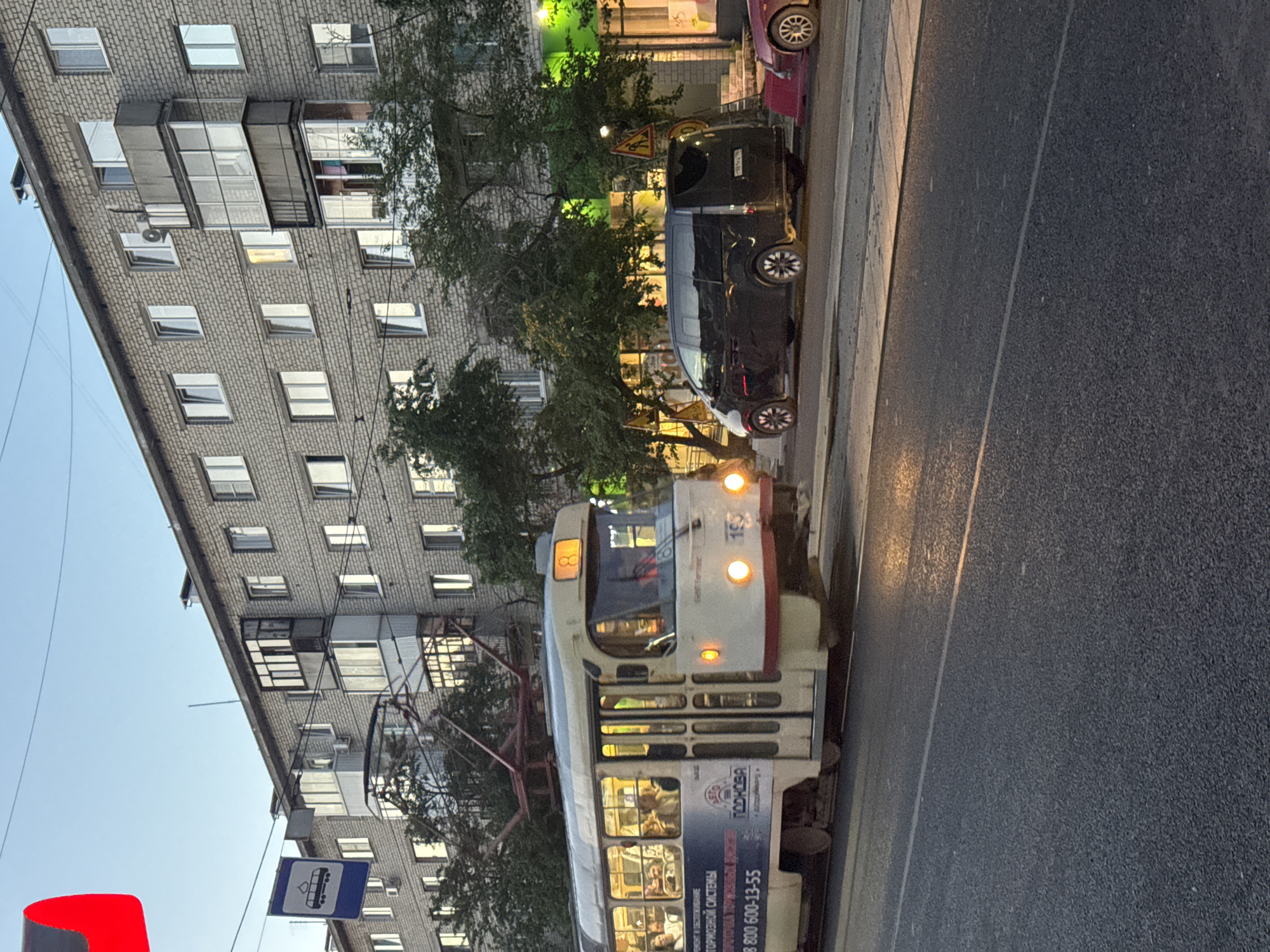 The image shows a street scene in Yekaterinburg, Russia, during what appears to be the late afternoon or early evening, given the lighting. In the foreground, a tram is traveling along its tracks on the left side of the road. The tram is white with red and blue accents, and it has several passengers visible through its windows. The tram has advertisements on its side, including one for "PODKOVA."
To the right of the tram, a dark-colored minivan is parked. Further to the right, a pink car is partially visible. The background features a multi-story brick apartment building with numerous windows and balconies. Green trees and foliage are present along the street, and there are overhead power lines, likely for the tram. A blue bus stop sign with a tram silhouette is visible on the left side of the frame, indicating a public transportation stop. The road surface is asphalt, and a white line marks the lane division.