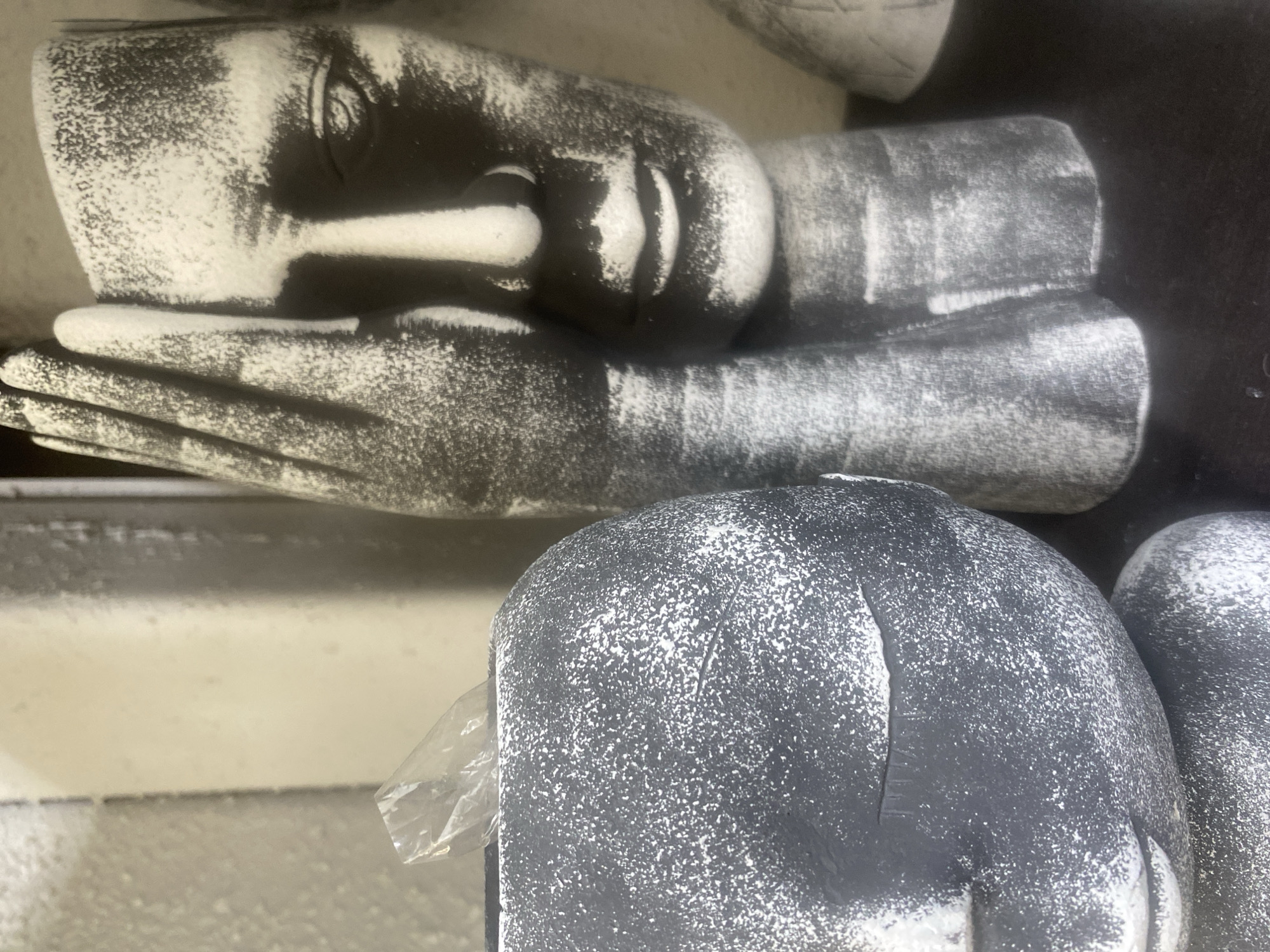The image shows a display of stone sculptures of reclining faces and heads. The sculptures appear to be dusted with a light coating of what might be paint or a similar substance, giving them a speckled appearance. The photograph was taken at the Yekaterinburg Stone-Cutting and Jewelry Art Factory Museum in Yekaterinburg, Russia.