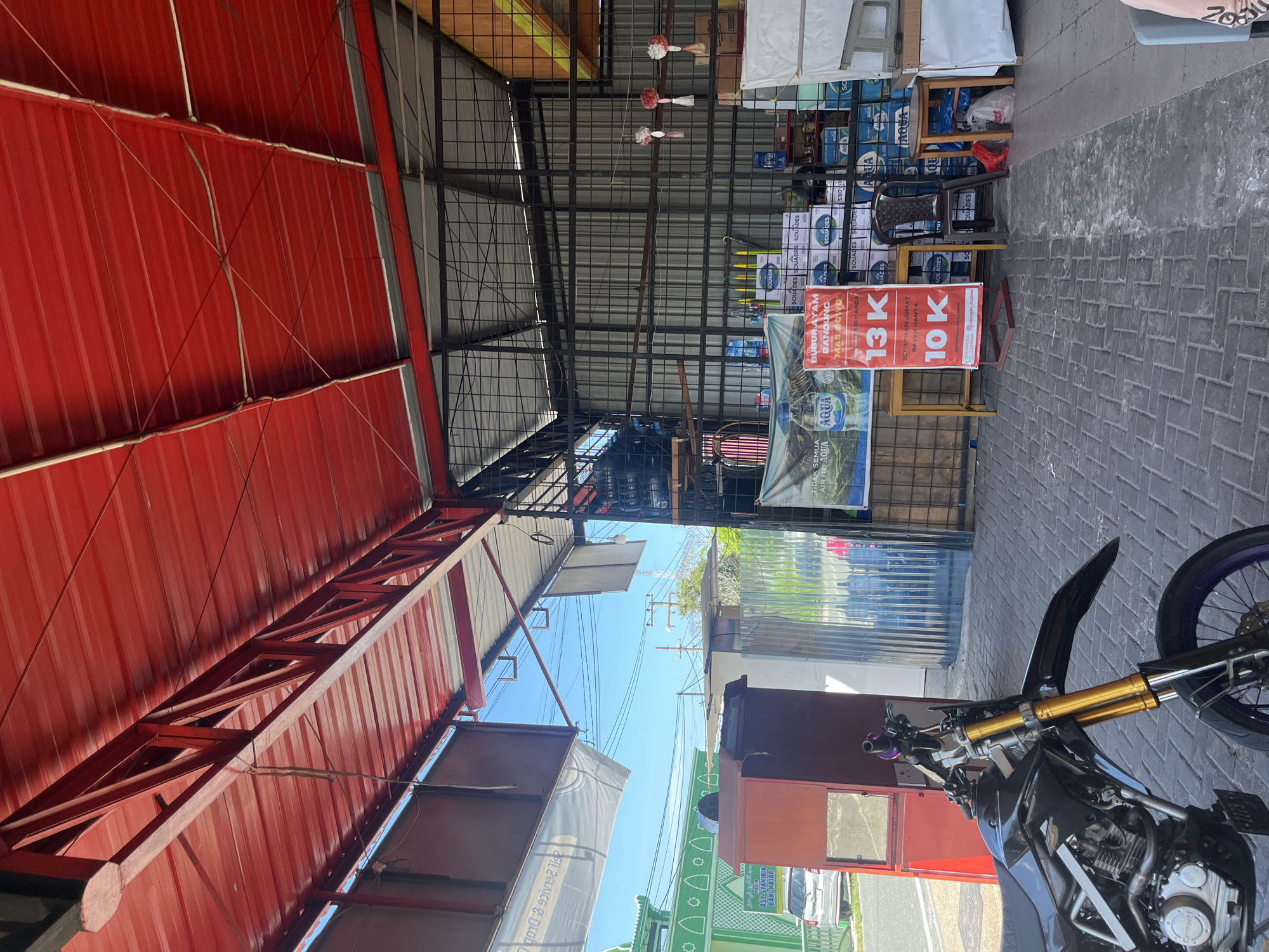 A motorcycle is parked in the foreground of an outdoor marketplace. The motorcycle is black with gold colored front suspension. Behind the motorcycle, to the left, is a red cabinet. To the right of the cabinet, there are stacks of bottled water and shelves. A metal frame structure supports a red roof overhead. In the background, a green building with a white patterned facade is visible. The ground is paved with brick. There are no people visible in the image.