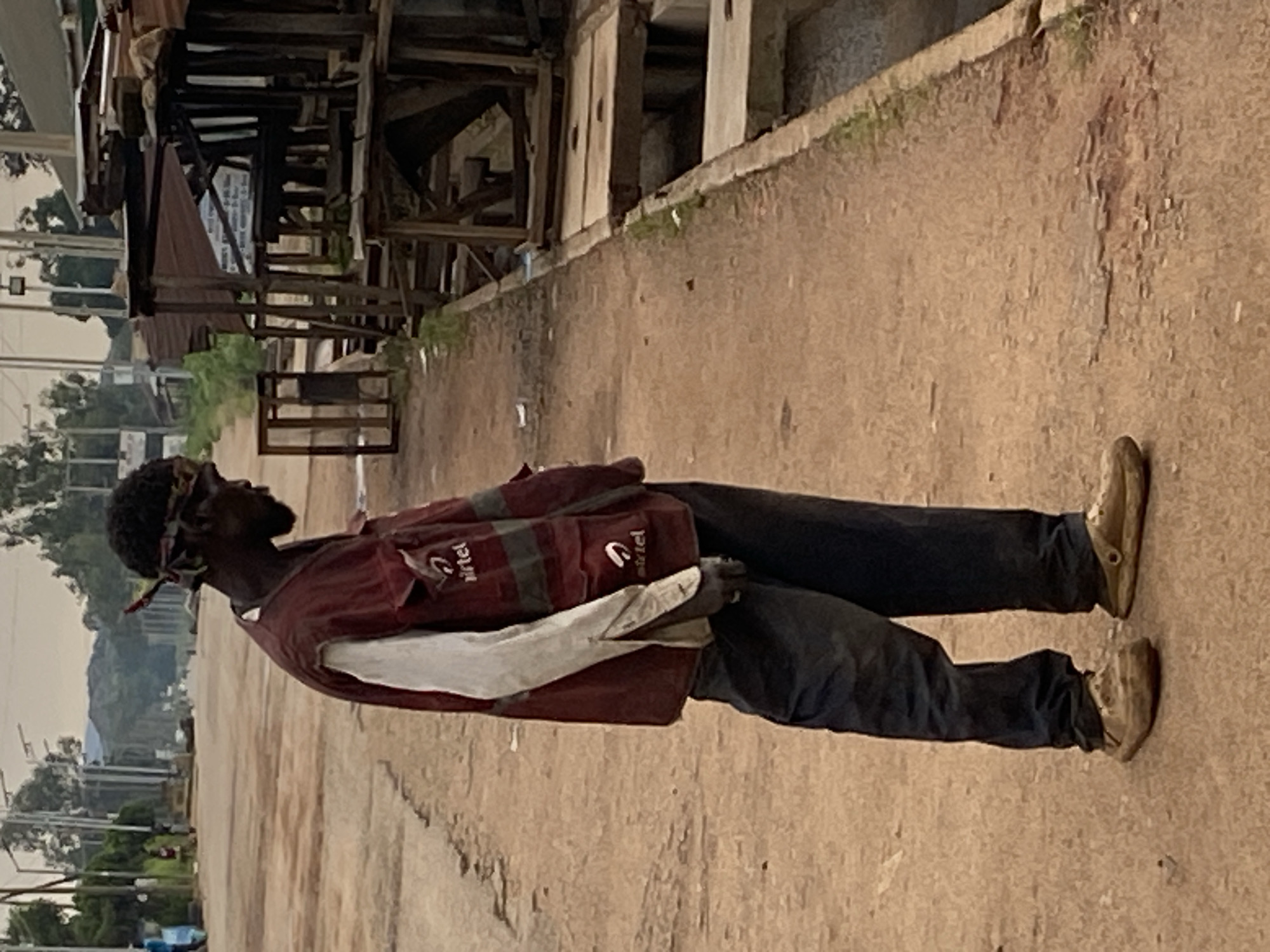 A side-view, medium shot captures a lone person standing on a dirt road in Vom, Nigeria. The individual, wearing a red vest over a white long-sleeved shirt and dark pants, has a bandana tied around their head. They are looking down and slightly to the right. The background reveals a street scene with wooden structures and vegetation, suggesting a market or informal settlement. A ditch runs along the right side of the road. The lighting indicates it is daytime.