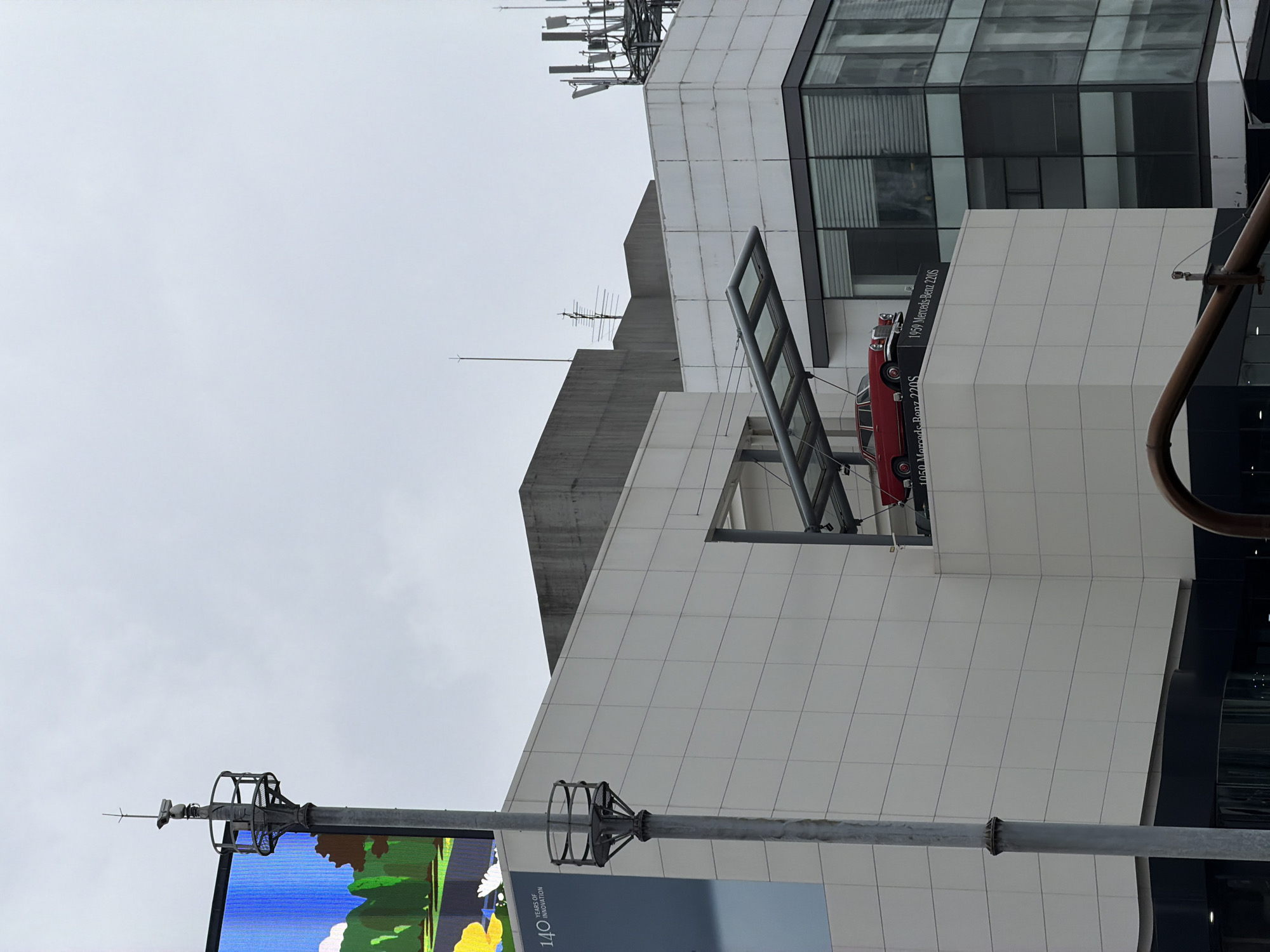 A red 1959 Mercedes-Benz 220S is displayed on a white pedestal on the exterior of a modern building in Seoul, South Korea. The car is positioned on a ledge projecting from the building, beneath a transparent canopy. The building features a grid of white panels on its facade, with a darker section at the bottom. To the left of the display, a tall pole with a camera and cage-like structure rises against a cloudy sky. Further left, a large digital billboard displays an abstract image with trees and a yellow element. The background shows a grey, overcast sky and other sections of the building with glass windows and communication antennas on the rooftop. A curved metal pipe is visible in the foreground. The text "1959 Mercedes-Benz 220S" is printed on the pedestal below the car.