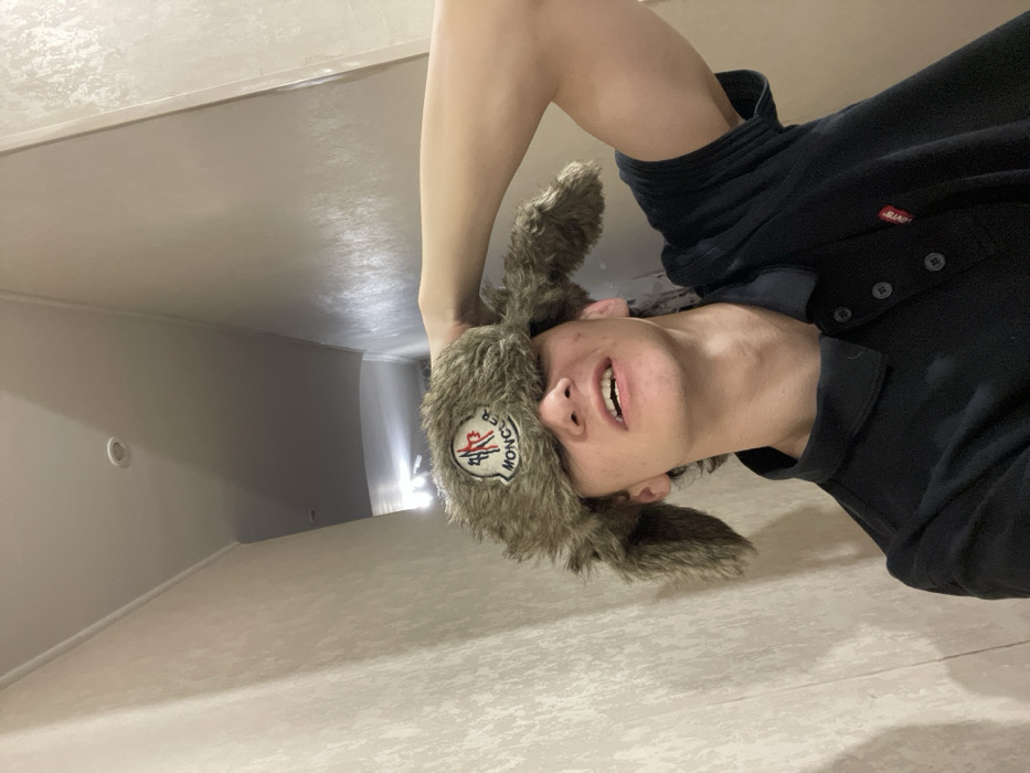 A person wears a fluffy Moncler hat and a black button-up shirt. The image is taken from a low angle, looking up at the individual, who appears to be positioned on a floor or similar surface. The background shows a room with light-colored walls and a hallway leading into the distance. The perspective is unusual, making the scene disorienting.