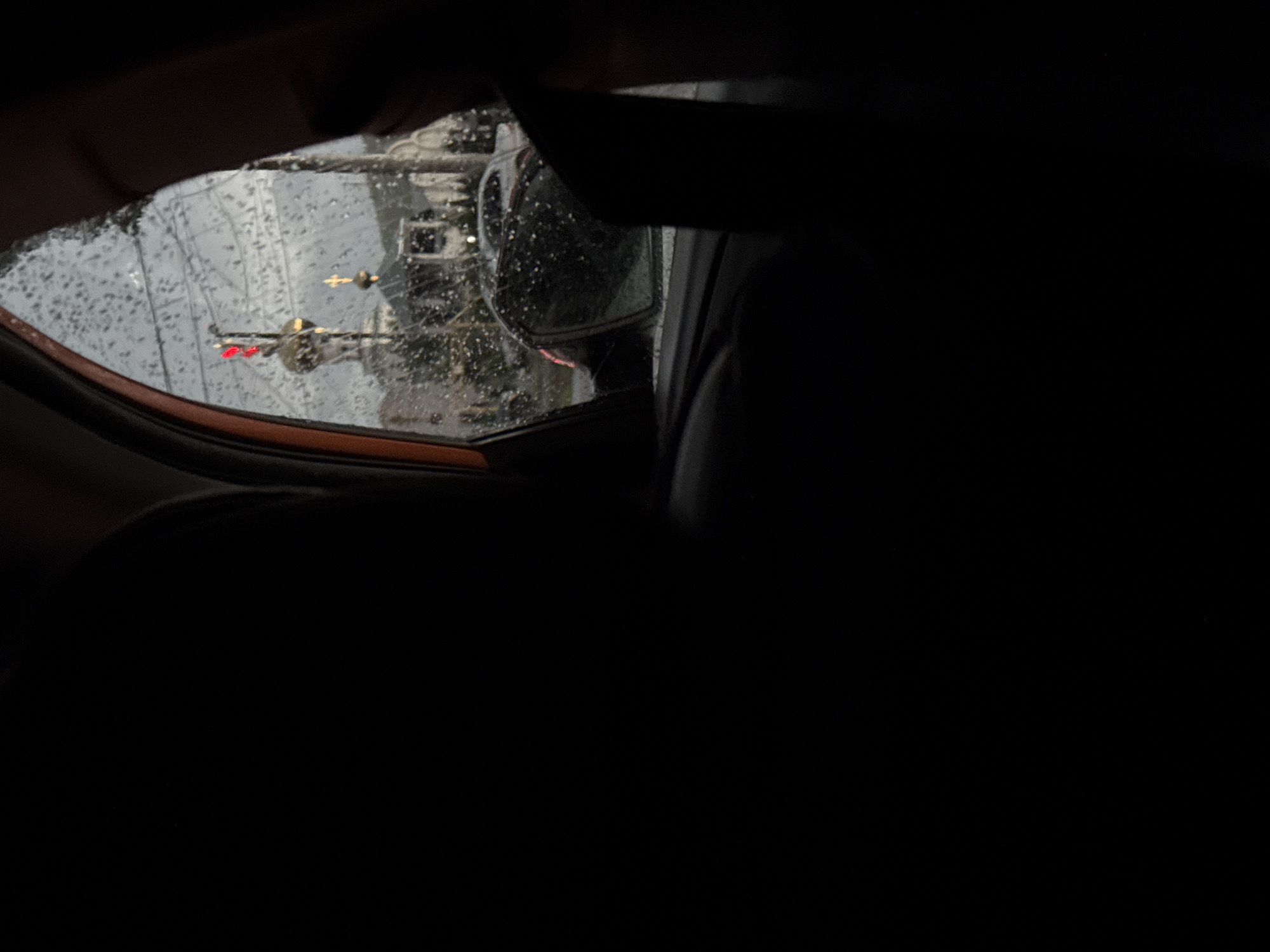 The view from inside a car on a rainy day shows a wing mirror reflecting a street scene. The mirror is covered in raindrops, obscuring the view, but a traffic light can be seen with a red light illuminated. Beyond the traffic light, a tall building is visible in the background. The majority of the image is dark, showing the interior of the car. The specific location within the city of Unknown is not discernible from the image.