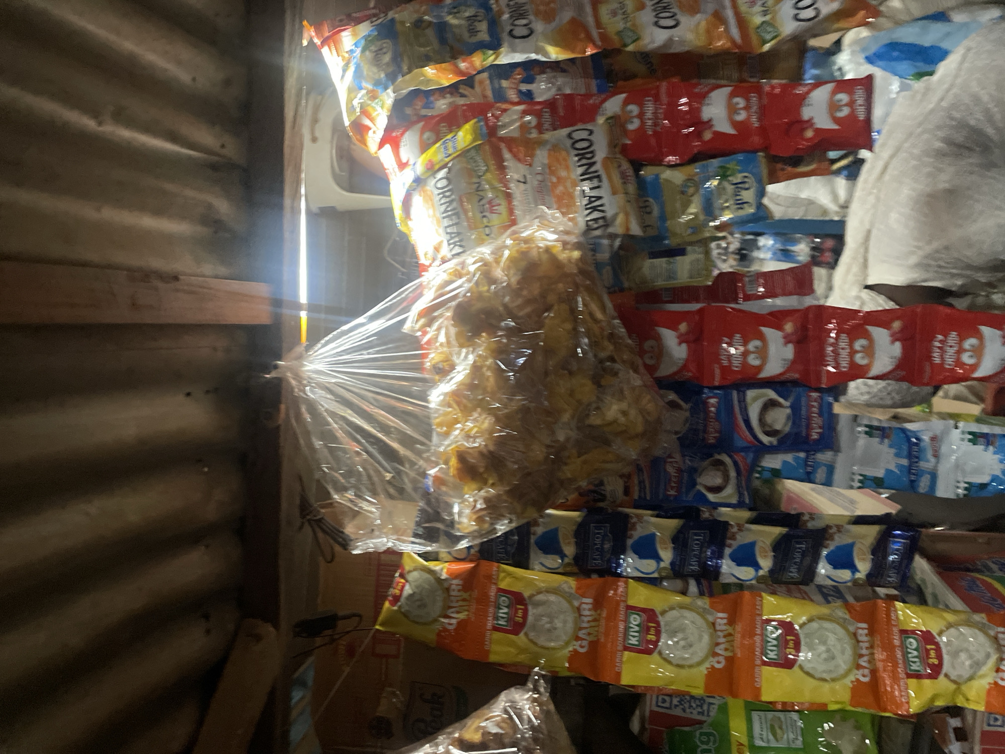 This image depicts a small shop in Sauka, Nigeria, likely a kiosk or a roadside stall. The setting is cramped, with shelves overflowing with various packaged goods, predominantly snacks and food items. The items are stacked high, creating a colorful and somewhat chaotic display.
Prominently visible are bags of "Cornflakes," "Happy Chinchin," and "Garri," which is a staple West African food. There are also packets of "Kivo" and "Topcafe," suggesting a mix of food and beverage products. The packaging displays bright colors and imagery, including cartoon characters on some of the "Chinchin" bags. The goods are arranged in rows, with some items, particularly a large bag of fried snacks, appearing to be loose in a plastic bag in the foreground.
The lighting suggests it might be daytime, with sunlight streaming in from the left, illuminating the products and casting some shadows. The wall to the left appears to be made of corrugated metal, a common building material for such small businesses in the region. The overall impression is one of abundance and everyday commerce in a local Nigerian setting. The image captures the vibrant nature of small-scale retail operations, offering a glimpse into the available consumer goods.
