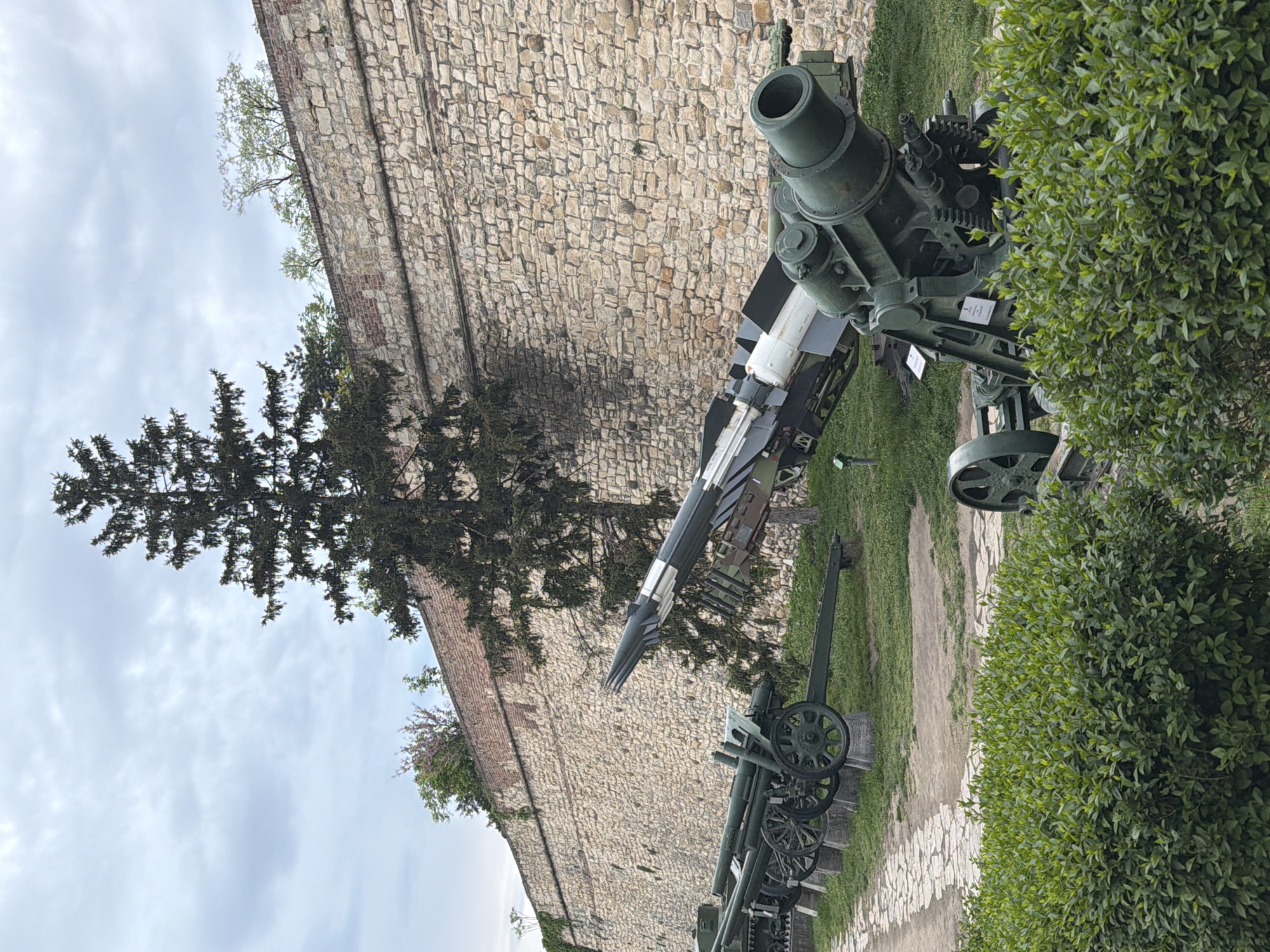 The image displays an outdoor exhibition of military ordnance, set against a historical stone fortress wall under an overcast sky. Given the context, this is likely part of the Military Museum located within the Belgrade Fortress (Kalemegdan) in Belgrade, Serbia.
In the background, a massive stone wall constructed from irregularly shaped light-colored blocks and some visible brick sections dominates the scene. Above the wall, a cloudy, light grey sky fills the upper portion of the frame. To the left, a tall, dark evergreen tree with dense foliage stands prominently, reaching towards the sky.
The foreground and mid-ground feature several pieces of green military equipment. On the right, a large, robust mortar with a wide, open muzzle is visible, positioned on a grassy patch. Adjacent to it and slightly to the left, a long, slender missile, dark with distinct white and black segments, rests on a launch platform. Further left and receding into the mid-ground, at least two more traditional artillery pieces, likely cannons or howitzers, are on display, featuring large spoked wheels. These are arranged on a combination of grass and a light-colored, paved pathway. A dense, green bush with small leaves occupies the far right, partially obscuring some of the equipment.
Small white informational signs are placed near some of the exhibits, but the text on them is not legible. The lighting suggests it is daytime, possibly early afternoon, with diffuse light due to the cloud cover. No people are visible, and no specific activity or event is taking place beyond the static display of historical military hardware.