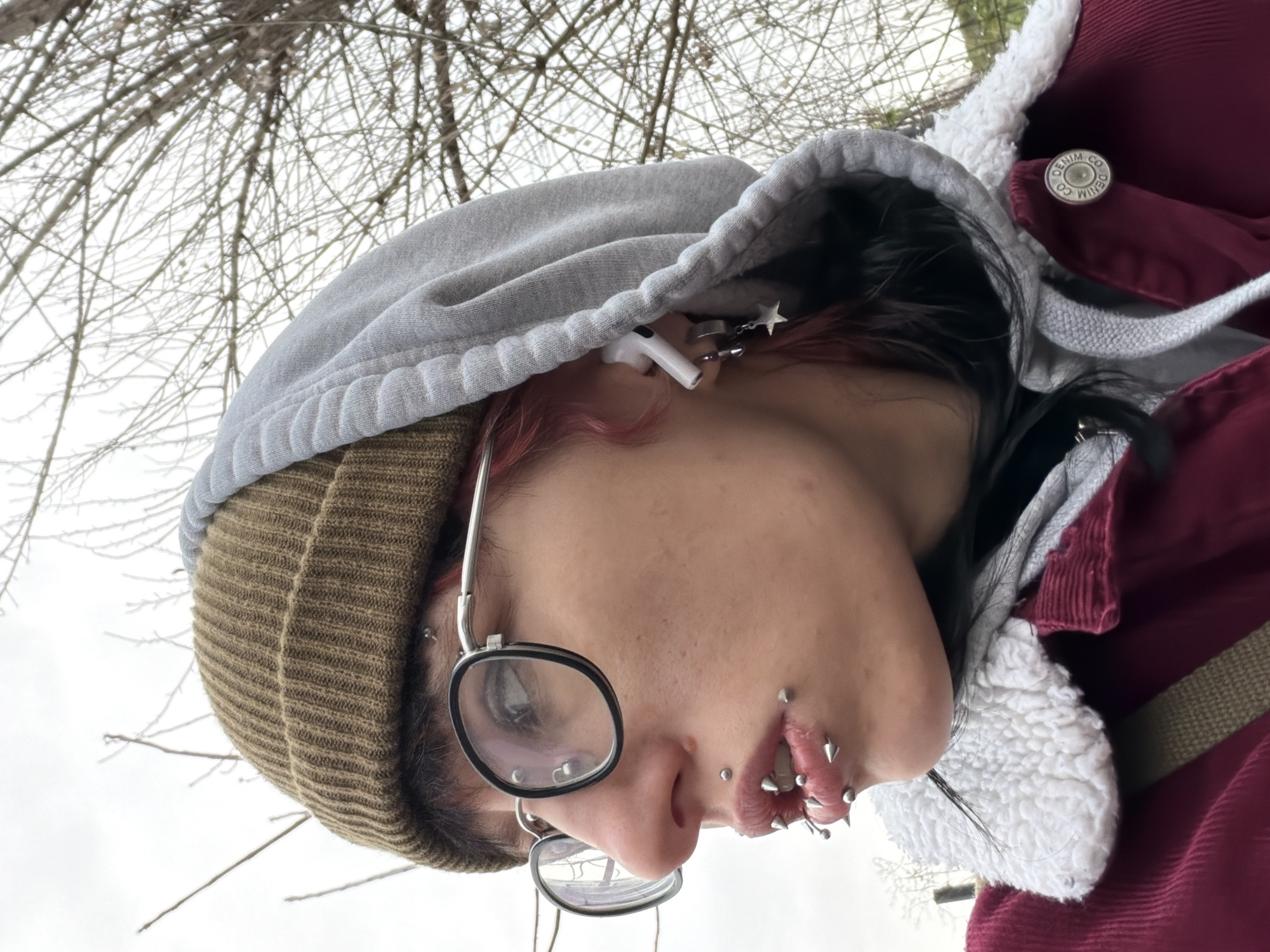 A close-up, low-angle photograph depicts a person from the chest up, outdoors against a backdrop of bare tree branches and overcast sky. The person, seen from the side, is wearing a brown knit beanie and a gray hoodie with a white faux fur-lined burgundy coat. They have round glasses, dark hair under the hood, and multiple piercings around their lips and face. An AirPod is visible in the left ear, adorned with a dangling star charm. The location is not identifiable as Sofia, Bulgaria based on the image alone.