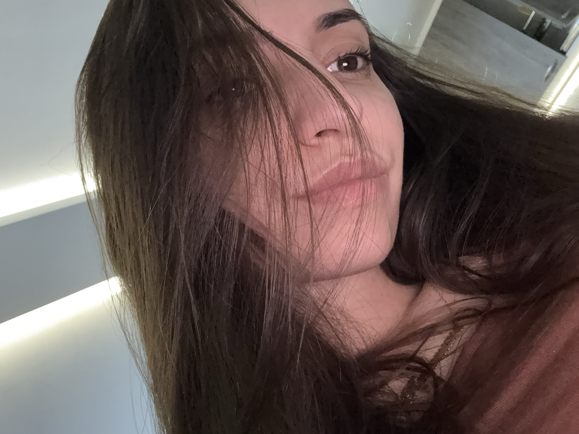 A close-up, low-angle selfie of a woman with long, dark brown hair partially covering her face. Her right eye is visible, and she has a neutral expression with slightly pursed lips. The background includes a bright, indirect light source, likely from overhead artificial lighting, and sections of a grey ceiling and wall. The location within the city of Plovdiv, Bulgaria is not discernible from the image provided.