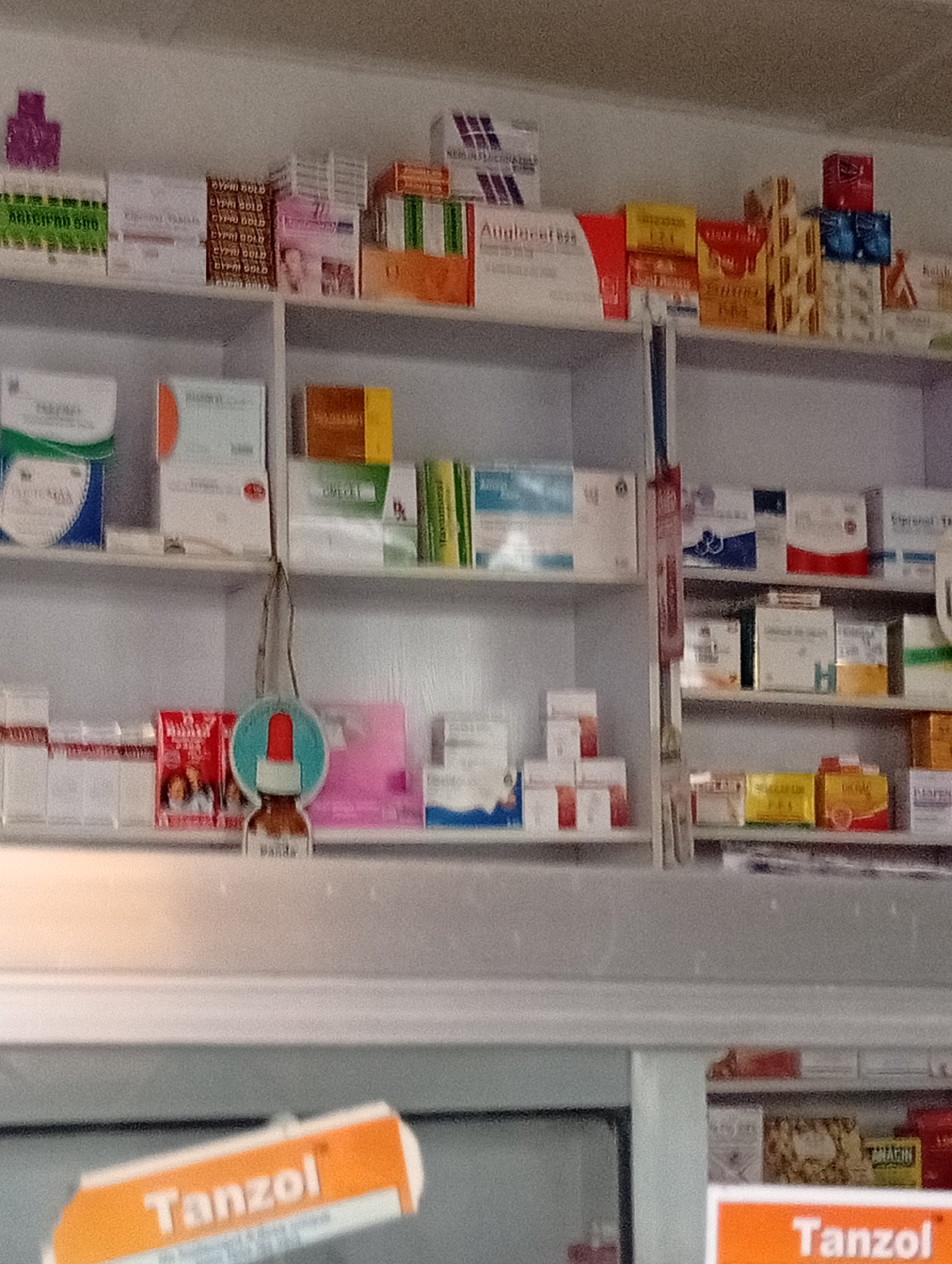 The image captures the interior of a pharmacy or drug store in Adankolo, Nigeria, displaying a wide array of packaged medications and health products on multi-tiered white shelves. No people are visible, indicating a static display of goods.
The shelves are densely packed with boxes of various sizes, shapes, and colors. On the top left shelf, green boxes labeled "ARICIPAD 500" are visible, alongside multiple gold-colored boxes stacked vertically, each bearing the text "CYPRO GOLD". Further to the right on the same shelf, a prominent red and white box clearly reads "Auglaclav 625". Other visible products on the upper shelves include boxes with partial labels like "NEUROBI...", "LIVINYA", and "CYPRO...".
The middle shelf features more neatly arranged boxes. A blue and white box on the left is labeled "PARASET", next to a white box with an orange stripe that might read "PASOCON". Green-packaged items are also present on this level. To the right, another section of shelves holds boxes with discernible letters such as "H" and "CYPROL TRA...".
In the foreground, a small, dark brown dropper bottle with a red top and blue label, marked "BANDA", hangs down, partially obscuring the lower shelves. The bottom shelf on the right displays boxes with texts like "DIVOC", "IYAFEN", and "AMAFIN". A large, orange and white sign is prominently visible at the very bottom of the frame, bearing the brand name "Tanzol". The overall scene suggests a well-stocked and organized pharmaceutical retail environment, brightly lit with artificial light.