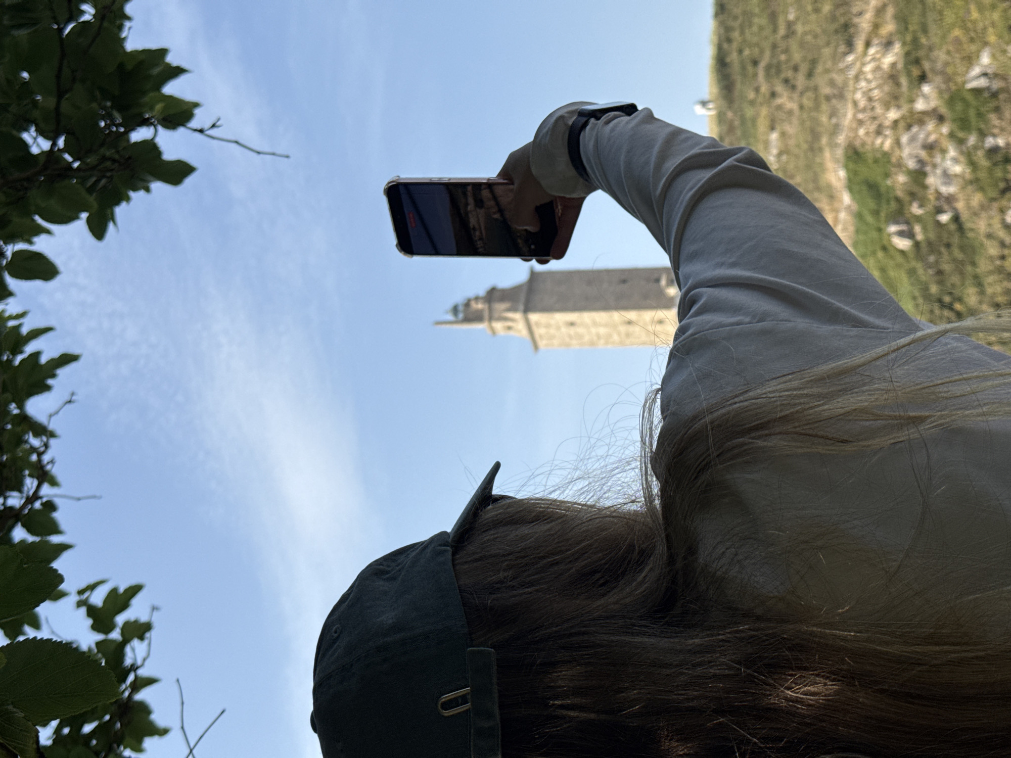 A person wearing a dark green baseball cap and a light-colored long-sleeved shirt is taking a photograph with a smartphone. The person is viewed from behind, with their long, blonde hair visible. The smartphone is held up in their right hand. In the background, the Tower of Hercules, a UNESCO World Heritage site and the oldest Roman lighthouse in the world, stands tall on a hill under a clear blue sky. The surrounding landscape is covered in dry grass and rocks. The image appears to be taken from a slightly elevated position, with tree leaves framing the top of the scene.