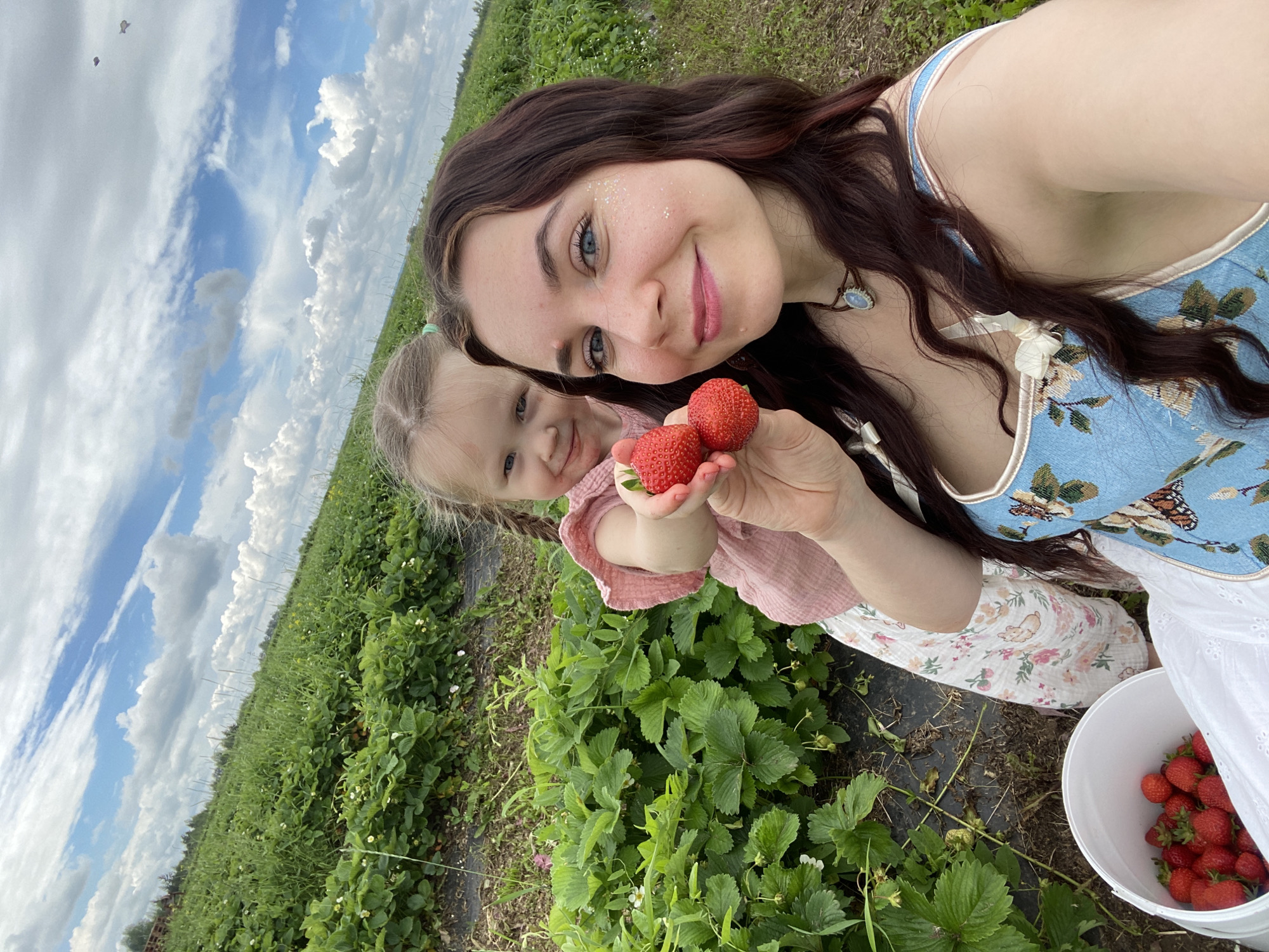 In the city of Foteni, Russia, a young girl and a woman are picking strawberries in a field. The girl, with blonde pigtails and a pink top, holds up two strawberries. The woman, with long dark hair, wears a blue floral dress and glitter on her cheek. A bucket of strawberries sits in the foreground. The field stretches into the distance under a partly cloudy sky.