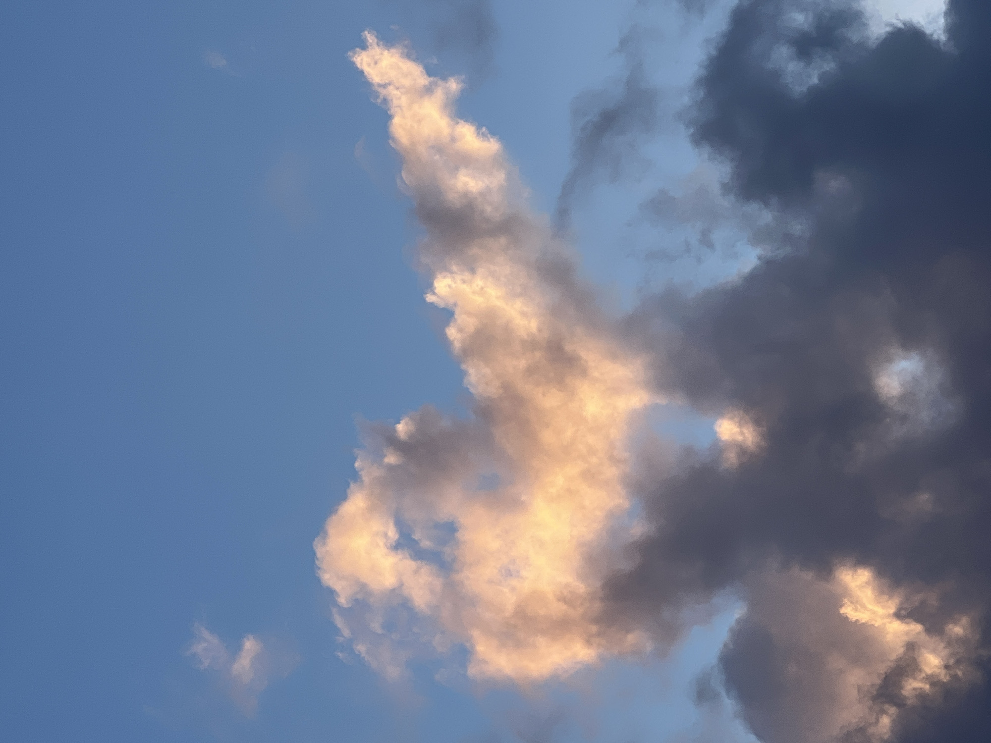 The image depicts a daytime sky with a mix of clouds, likely taken in Chaikovsky, Russia. The sky is a gradient of blue, with the lightest blue in the upper left and a slightly darker blue mixed with hints of gray towards the right. The clouds are of varying density and color. One prominent cloud formation in the center is illuminated with a warm, golden light, contrasting with darker, gray storm clouds to the right. The sun is likely setting or rising, casting a golden glow on the clouds.