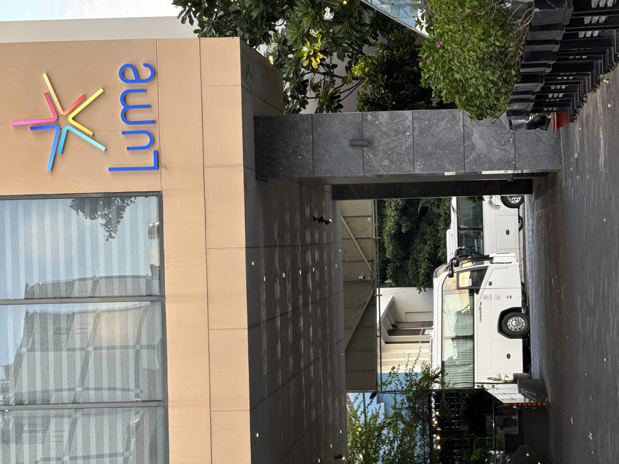The image depicts the exterior of a building with a prominent "Lume" logo and a stylized star design. The building has large windows with horizontal blinds. A covered walkway extends from the building, supported by dark gray, marbled pillars. Two white buses are parked beneath the walkway, with their doors open on one of the buses. Lush green trees and plants are visible around the building and the buses. The ground is paved. The scene is likely a hotel or a similar establishment in Nha Trang, Vietnam, given the architecture and the presence of buses, suggesting transportation services.