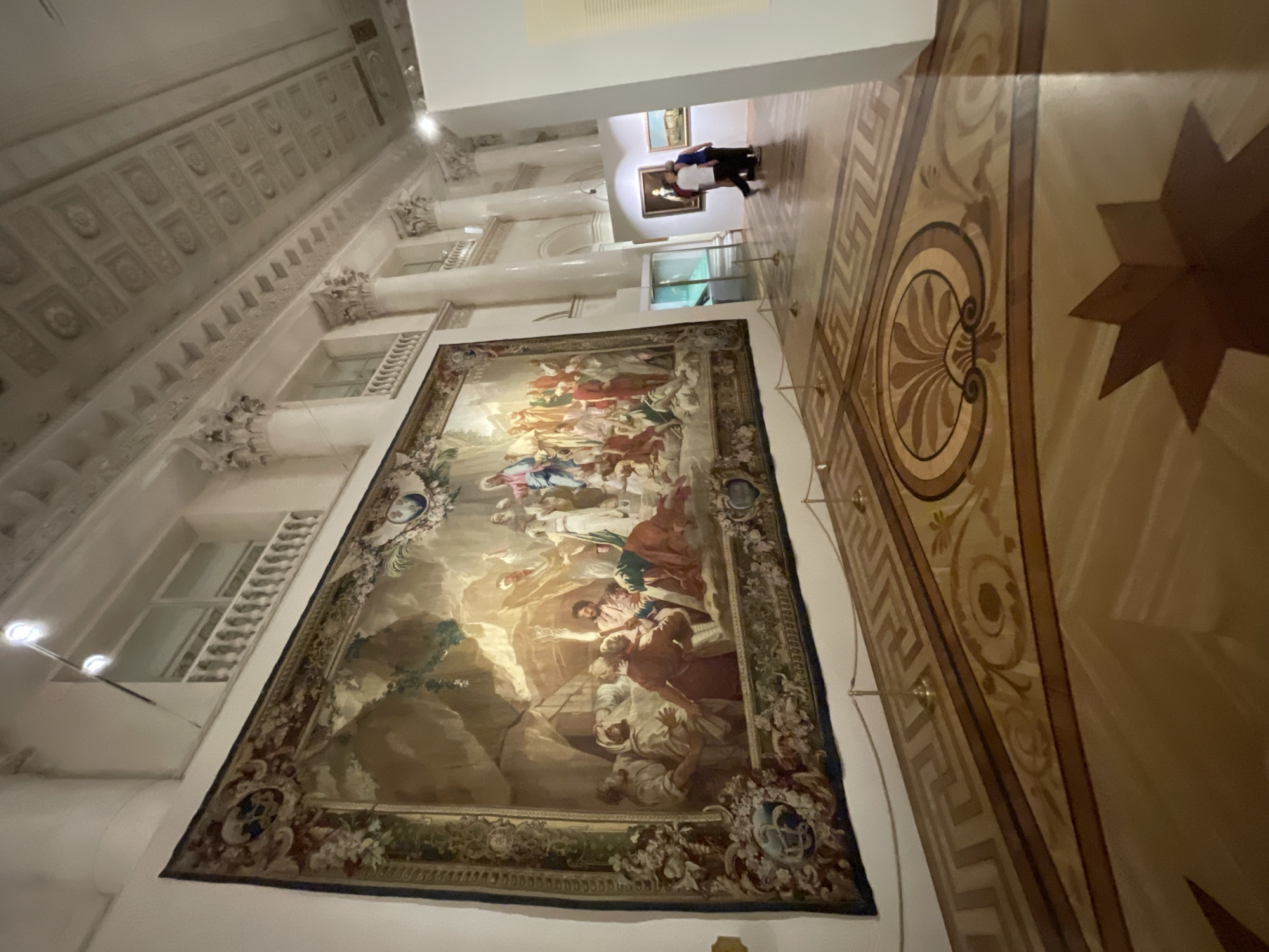 This is an indoor image of a gallery or museum in Saint Petersburg, Russia, likely within the State Hermitage Museum or the Russian Museum due to the opulent architecture and vast collection. The focal point is a large, richly detailed tapestry hanging on a white wall. The tapestry depicts a religious scene with numerous figures in classical attire, rendered in muted, earthy tones with areas of bright illumination suggesting a dramatic narrative. The tapestry is bordered by intricate floral and heraldic designs.
In the foreground, a highly polished wooden floor is visible, featuring an elaborate parquet design with geometric patterns and decorative inlays, including a large starburst motif and a Greek key border. Stanchions with ropes guide visitors through the space, indicating a controlled viewing environment.
In the background, the room opens up to reveal more of the museum's architecture, characterized by tall, fluted columns, ornate ceiling moldings, and large windows with balustrades. Several other artworks, including framed paintings, are displayed on the walls in the distance, and a few visitors are seen walking through the exhibition space. The lighting appears to be a mix of ambient and directed spotlights, common in museum settings to illuminate the artwork. The overall impression is one of a grand and culturally significant institution.
