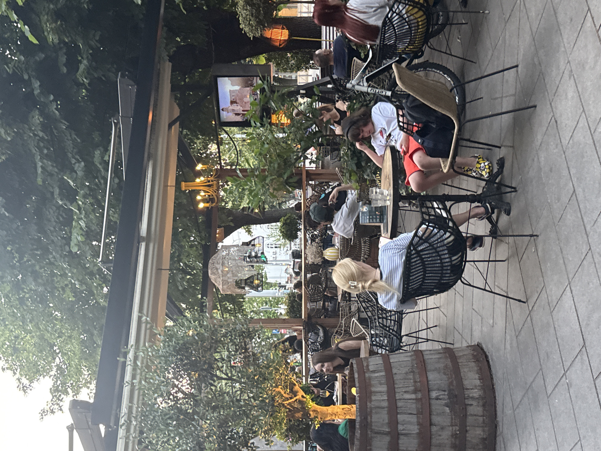 The image depicts an outdoor seating area of a restaurant or cafe in Belgrade, Serbia, likely during the late afternoon or early evening. The scene is set amidst lush greenery and trees, providing shade from the sun. Several patrons are seated at tables, engaged in conversation or relaxing.
In the foreground, a woman with blonde hair pulled back is seated at a table, facing away from the camera. She is wearing a striped shirt and has a laptop open in front of her. To her left, a large wooden barrel serves as a decorative element. Another table to the right has a younger individual with dark hair wearing a white t-shirt and red shorts, looking down at their phone. They are seated on a woven chair with a bicycle leaning against it.
Further back, other individuals are visible at various tables, some interacting with each other, while others appear to be dining or enjoying drinks. The restaurant features a covered outdoor area with modern lighting fixtures, including a chandelier. A television screen is mounted overhead, displaying an image of a building. The ground is paved with interlocking tiles. The overall atmosphere suggests a relaxed urban dining experience.