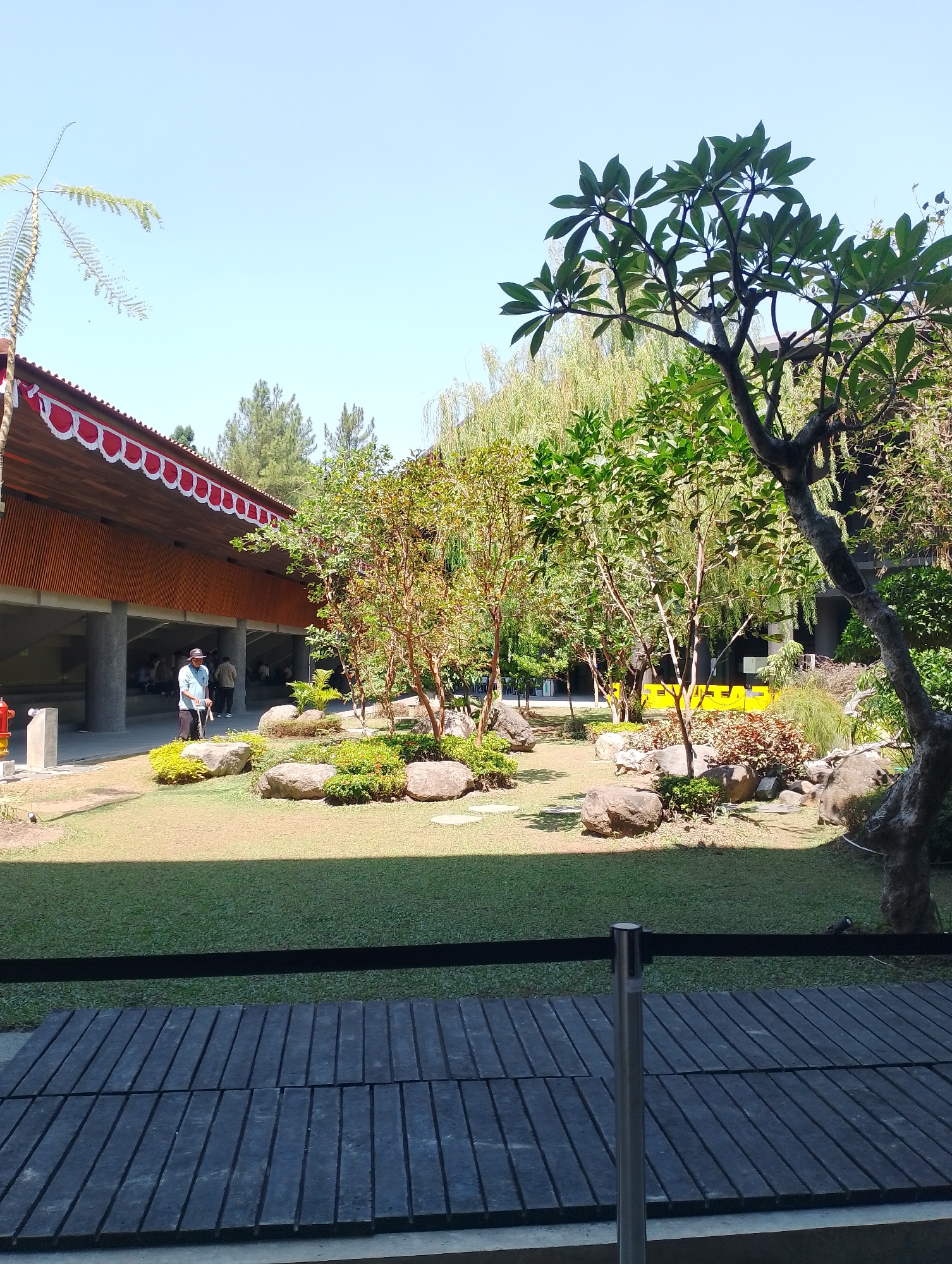 The image depicts an outdoor scene with a modern building featuring a red and white decorative awning. In front of the building is a lush garden with manicured grass, large rocks, and several trees. A person wearing a light blue shirt and a hat is walking with a cane in the garden. Other people are visible in the background, some sitting and others walking. The foreground shows a wooden walkway with dark planking and a metal stanchion with a black ribbon. In the background, behind the garden, there's a bright yellow structure with text. The sky is clear and blue. This scene is located at the Museum of Indonesian Computer History in Depok, Indonesia.