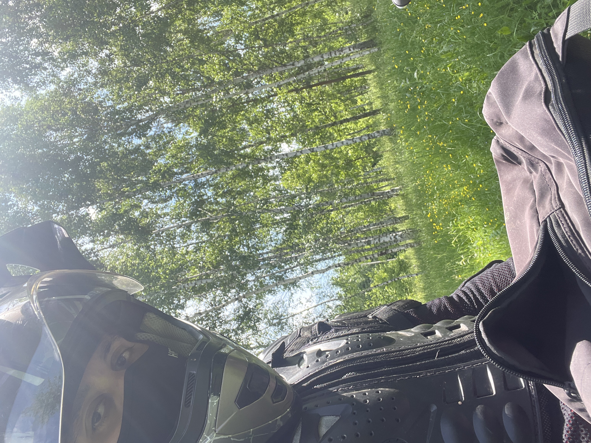 A person wearing a motorcycle helmet, face mask, and protective gear is positioned in the foreground of the image. The person's eyes are visible through the helmet's visor. A black bag with an open zipper is in the lower part of the frame. Behind the person, a lush green meadow dotted with small yellow flowers stretches towards a dense forest of birch trees with white trunks. The sky above is bright and sunny. The scene suggests an outdoor activity, possibly on a motorcycle, in a natural environment that could be the outskirts of Vasilyevo, Russia, given the characteristic birch trees.