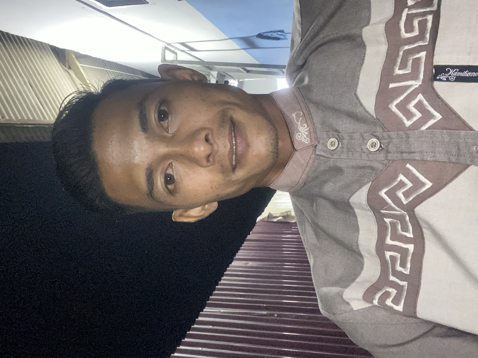 The image presents a close-up, rotated portrait of a man, likely a selfie, in Seuneubok Teungoh, Indonesia. He has short, dark hair, dark eyes, and a faint mustache and beard stubble. A slight, friendly smile is visible on his face, and his skin appears somewhat shiny, possibly indicating sweat or a warm environment.
The man is wearing a short-sleeved, collared shirt in shades of light brown and white. The shirt features a distinctive pattern with broad vertical stripes and an intricate geometric design in white and a darker brown. A small, dark blue label with the brand name "Kamiliano" in white script is visible on the right chest area of the shirt. The collar also has a delicate white embroidered motif.
The background is varied, suggesting a semi-enclosed or indoor setting. To the man's left (when oriented correctly), there's a bright overhead structure, possibly a ceiling or awning, with visible light fixtures and a patch of light blue and white surface above. To his right, a dark, textured area forms part of the background. Below him, a corrugated reddish-brown metal surface, consistent with a roof or wall, is prominent, showing parallel ridges and reflections of light. The overall scene suggests a casual environment. The activity is the man posing for the camera.