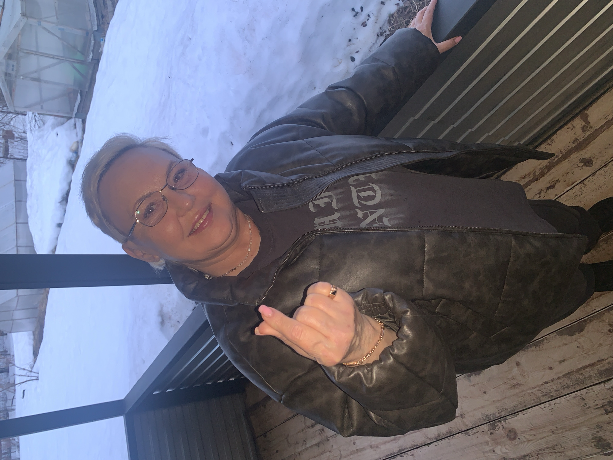 A woman wearing glasses, a necklace, and a brown leather jacket over a dark t-shirt is seen posing for a photograph. She is extending her hand and middle finger towards the camera. She is wearing a gold bracelet and a ring on that hand. The background reveals a snowy landscape with what appears to be a greenhouse or shed. The scene is likely taken from a balcony or porch with wood planks visible. The location appears to be Chaikovsky, Russia due to the background landscape and the person's attire, which are typical of that region.