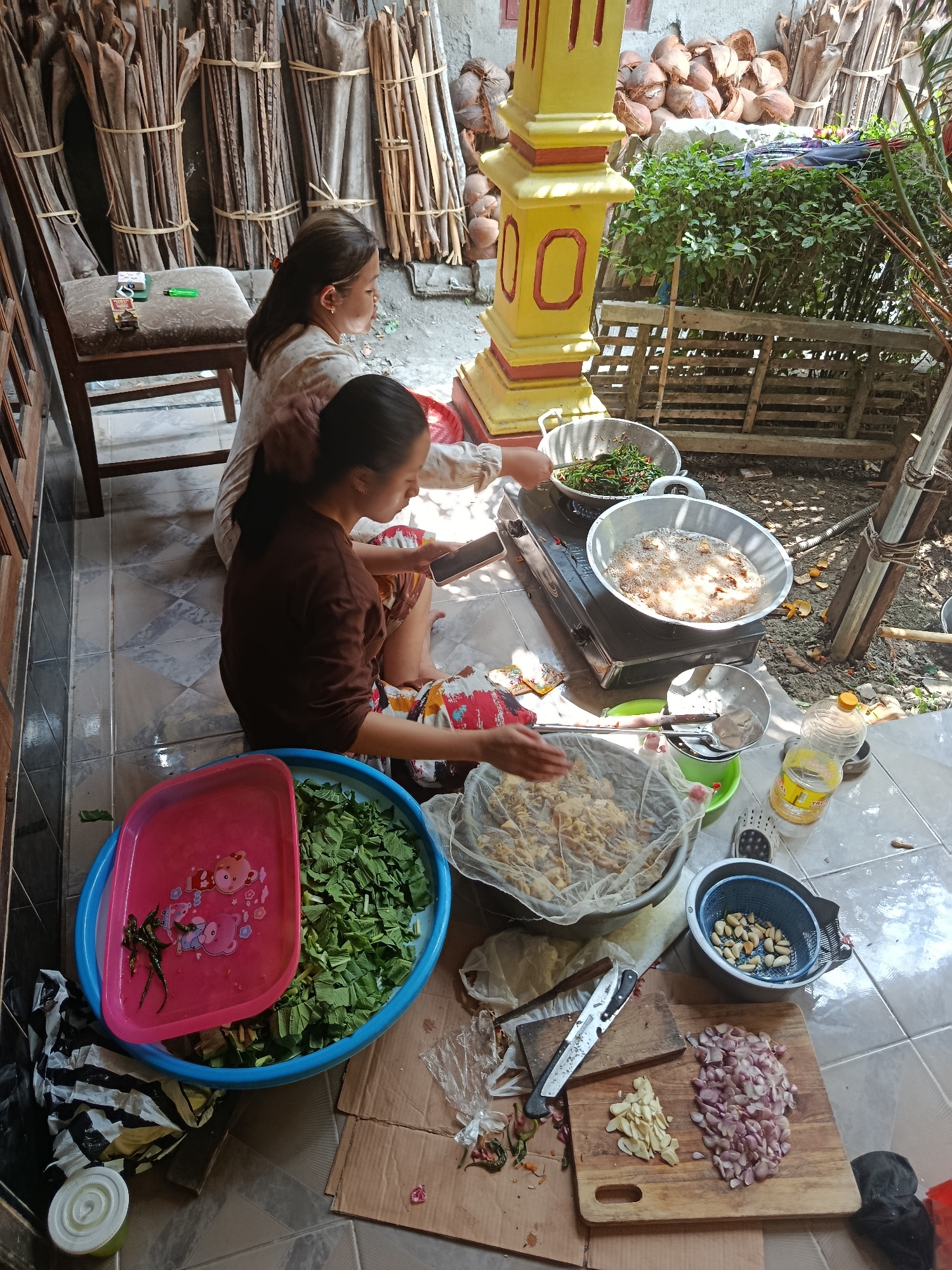In Sarwadadi, Indonesia, two women are engaged in food preparation outdoors, likely in a residential courtyard, given the presence of a tiled floor and a partially visible wall. The scene is cluttered with cooking implements and ingredients, suggesting an active and somewhat informal culinary environment.
In the foreground, a wooden cutting board displays a pile of thinly sliced red onions and a smaller pile of sliced garlic. Next to the cutting board, a blue colander holds a quantity of shelled peanuts, and a metal strainer with a handle rests nearby. A large blue plastic basin is filled with chopped green leafy vegetables. A pink plastic tray, decorated with cartoon bears, sits atop the basin.
To the right, a portable gas stove is in operation. Two large metal pans are on the stove. The pan on the left contains green vegetables and chili peppers being stir-fried. The pan on the right holds a significant amount of oil in which items are being deep-fried, evidenced by the bubbling and splattering oil. A clear plastic bottle of cooking oil stands near the stove.
One woman, wearing a brown long-sleeved shirt and patterned shorts, is seated on the floor and appears to be using a mobile phone. The second woman, wearing a cream-colored top, is also seated and is actively stirring the contents of the stir-fry pan with a utensil.
In the background, bundles of dried plant stalks are stacked vertically, tied with string, and leaning against a wall. A yellow and red pillar, possibly part of a building's decorative structure, stands to the right of the stalks. A pile of coconut shells is visible behind the pillar. A wooden chair with a cushioned seat is partially visible in the upper left corner, with a pack of cigarettes and a green lighter resting on it. The overall lighting suggests it is daytime, with strong sunlight casting shadows. The ground is a mix of tiled flooring and dirt.