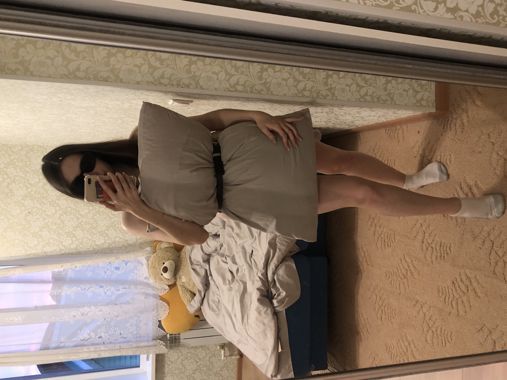 This mirror selfie captures a person standing in what appears to be a bedroom in Noviy, Russia. The subject, likely female, has long dark hair and wears dark sunglasses. They are holding a light grey pillow to cover their torso, with their left hand holding a light-colored smartphone to take the picture, and their right hand resting on the pillow. The person is wearing white ankle socks.
The room's interior, as reflected in the full-length mirror, features patterned wallpaper with a creamy background and delicate, light-colored floral or damask designs. The floor is covered with a plush, light brown carpet that has a textured, abstract pattern. To the side, a bed is visible with grey bedding and a dark blue frame. On or near the bed, a light brown teddy bear and a yellow stuffed animal or pillow can be seen. A window with white lace curtains, also featuring floral patterns, allows natural light into the room, and part of a heating unit or radiator is visible below it. The setting conveys a casual and private domestic space. No visible text is present in the image. The person's pose with the pillow suggests participation in a popular social media trend.