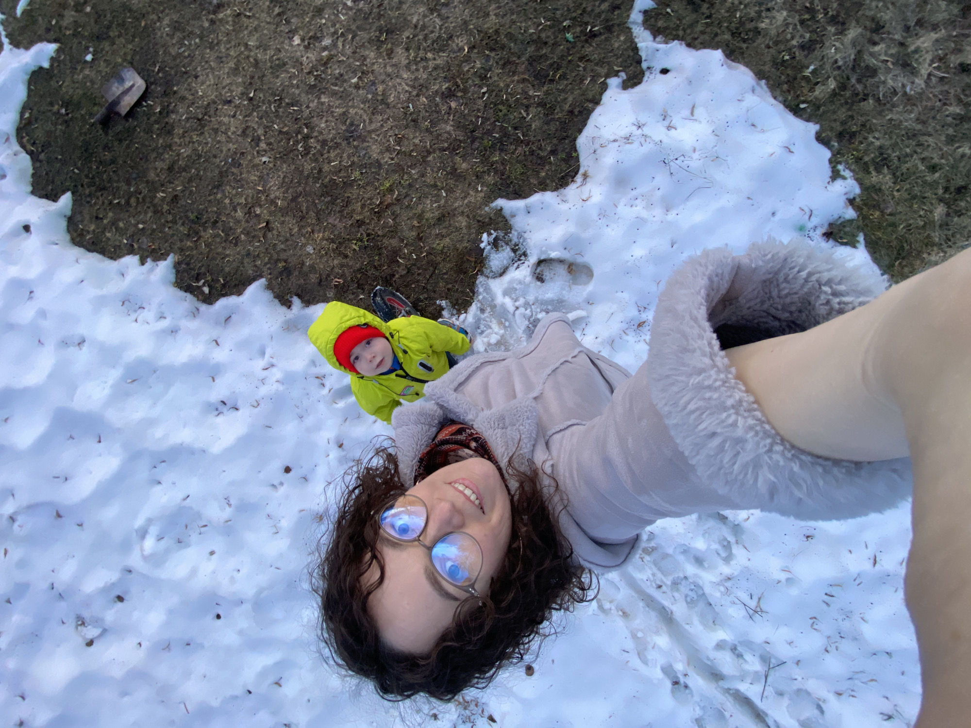 The image is a selfie taken in an outdoor area with patches of snow and grass. The photographer, a woman with curly dark hair and glasses, is looking up at the camera and smiling. She is wearing a light-colored coat with fur trim. To her left and slightly behind her, a young child wearing a bright yellow jacket and a red hat is also looking up at the camera. The ground is a mix of snow-covered areas and patches of grass. A small shovel is lying on the grassy part of the ground. Ольховка, Russia, is not confirmed in this image.