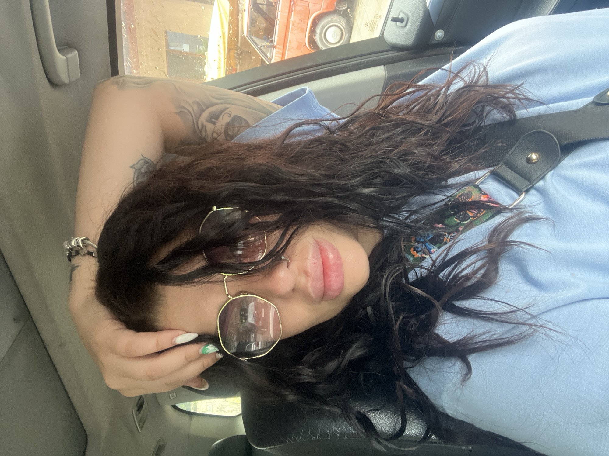 The image shows a young person, likely female, reclining inside a vehicle, captured from a tilted, slightly overhead perspective. She has long, dark, wavy hair that partially covers her face and shoulders. She wears large, roundish-rectangular sunglasses with gold frames, obscuring her eyes, and a light blue short-sleeved top. Her lips are full, and a small nose piercing is visible.
Notable accessories include a silver-colored bracelet on her left wrist and fingernails painted white, with one visible nail featuring a green abstract design. Intricate tattoos are visible on her left arm and shoulder, depicting what appears to be a face or figure amidst swirling patterns. A black strap, possibly from a bag or seatbelt, with gold hardware, is seen across her chest.
The setting is inside a car, with a black seat visible and grey interior elements like the headliner and grab handle. Through the window, which is covered in rain droplets suggesting wet weather, parts of the outside world are visible. An orange vehicle, possibly a truck, and some yellow elements are discernible in the background. The scene is set during daytime in Plovdiv, Bulgaria. The person's pose appears relaxed and confident. No visible text is present.