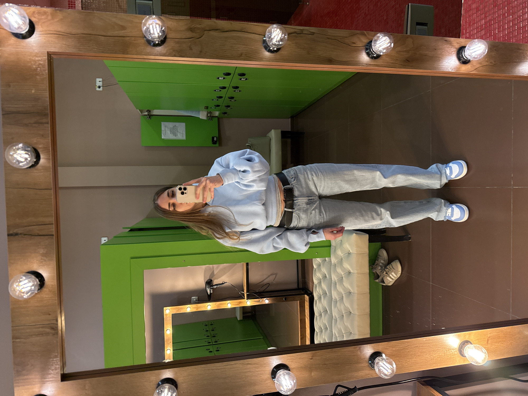 A young woman is captured taking a mirror selfie in a well-lit indoor setting, likely a changing room or dressing area. She is reflected in a large, vertically oriented mirror that features a thick, light-colored wooden frame adorned with multiple clear, vintage-style light bulbs along its perimeter.
The woman has long, light brown hair and is dressed in a light blue long-sleeved top, light-wash jeans, a black belt, and light blue and white sneakers. She holds a light-colored smartphone up, presumably an iPhone, to photograph her reflection.
Within the mirror's reflection, the background reveals a row of vibrant green lockers, strongly suggesting a locker room or gym environment. To the woman's right (viewer's left in the reflection), a light-colored, tufted bench or ottoman is visible. Further in the reflection, another, smaller rectangular mirror, also framed with light bulbs, sits on a dark surface. On the dark floor, near the tufted bench, a pair of grey sneakers with white stripes can be seen. A small white rectangular sign with illegible text is attached to one of the green locker doors.
The room where the main mirror is located has a dark brown floor and a section of red-tiled wall visible in the upper right. Electrical wiring and an outlet are also visible on the wall behind the mirror frame. The scene is brightly illuminated by the integrated lights of both mirrors, creating a clear and focused view of the woman and her surroundings.