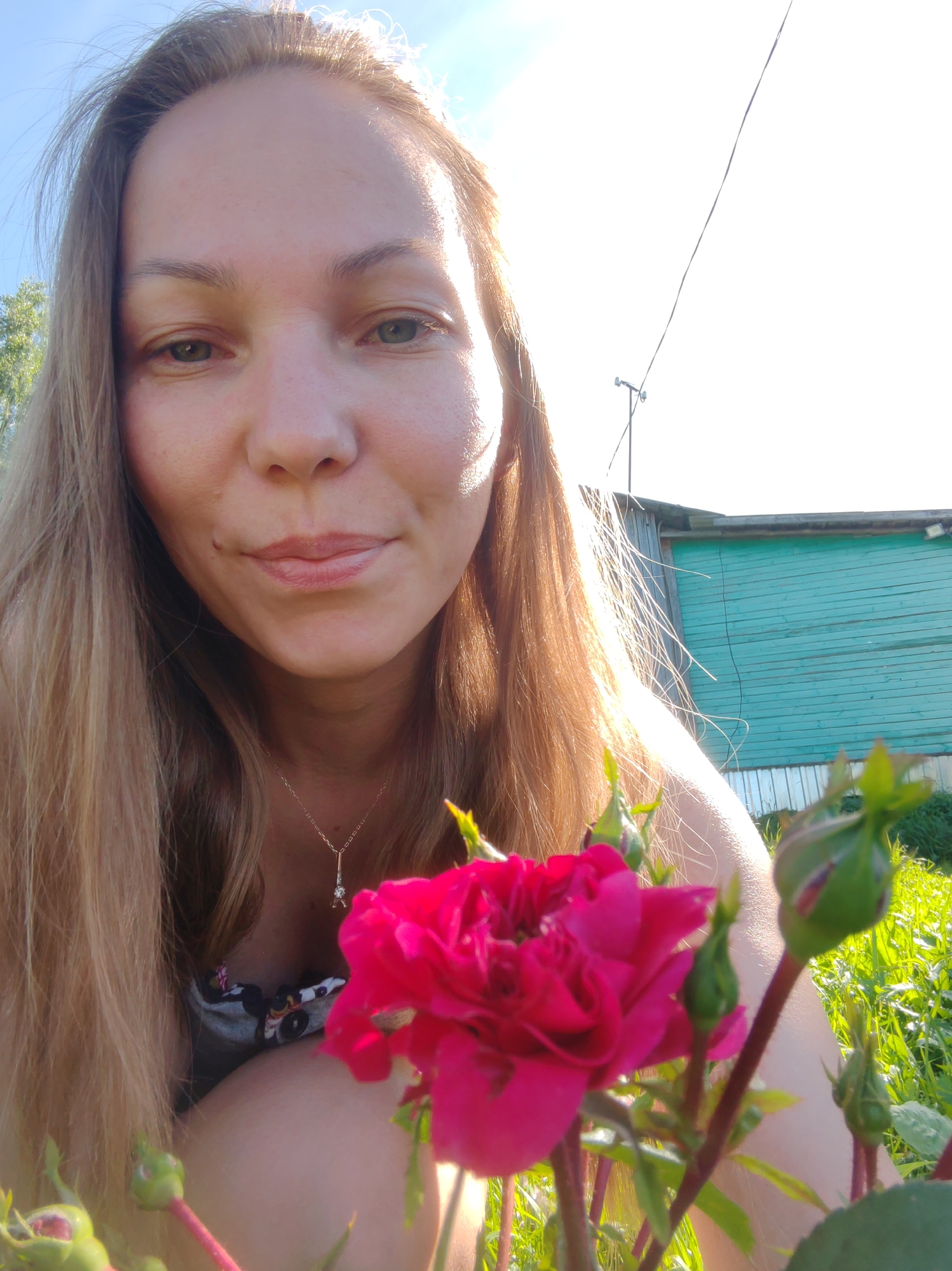 The image is a selfie taken outdoors, likely in a rural or suburban setting, given the presence of a wooden building with a turquoise facade, grass, and vegetation. A woman with long, light brown hair is the primary subject. She is smiling subtly and looking towards the camera. A bright pink rose is prominently featured in the foreground, partially obscuring her face and shoulder. The background shows a bright sky, the aforementioned turquoise wooden building, and some green foliage. The lighting suggests it is daytime, possibly late afternoon due to the warm glow. The overall impression is a casual, nature-focused portrait. The city of Чухлэм, Russia is not explicitly identifiable within the image itself; the context of its location is provided as external information.