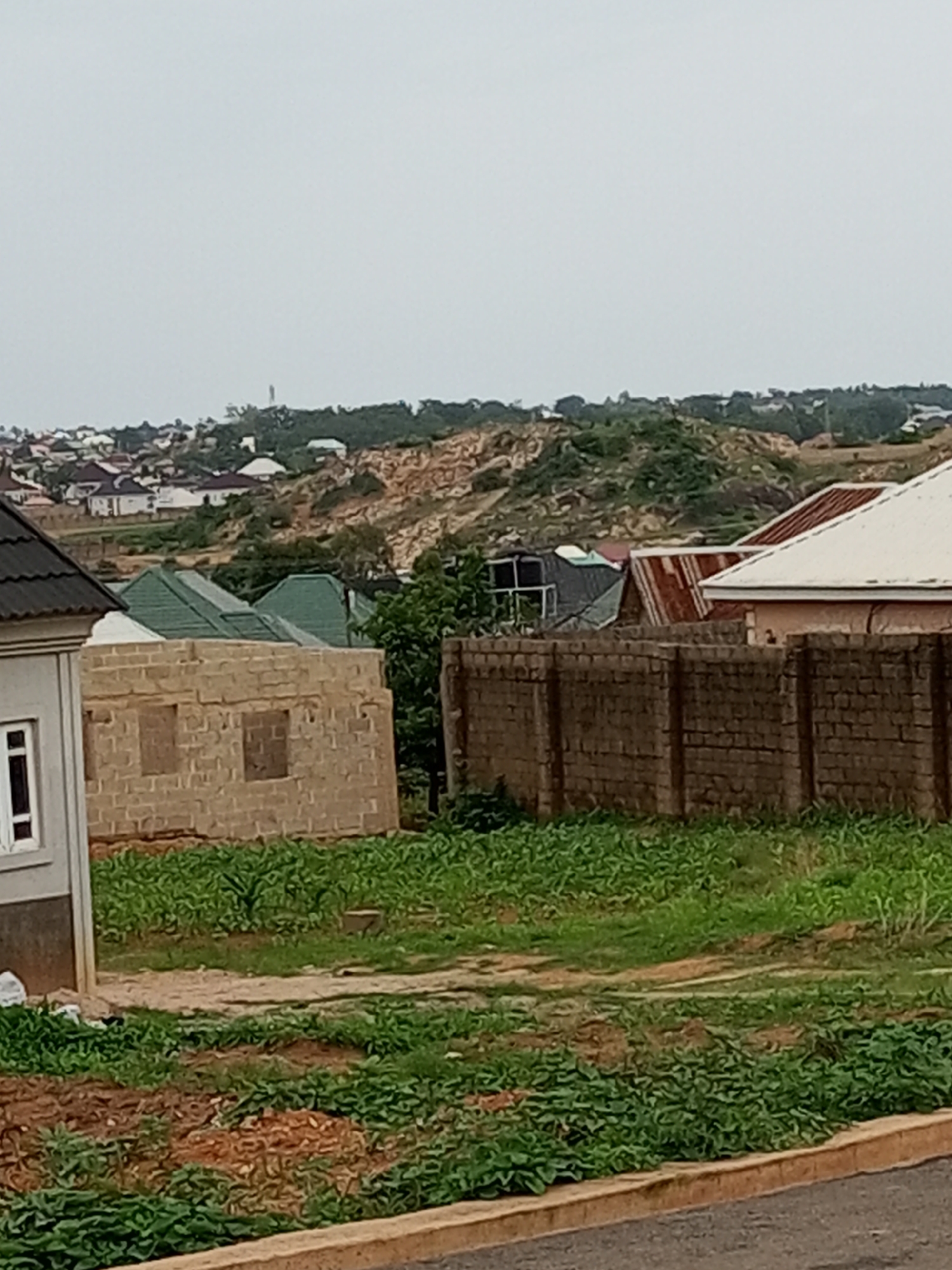 The image depicts a partially constructed residential area in Gura Top, Nigeria. In the foreground, a building with exposed brick walls and two window openings is under construction. To its left, a finished house with a dark pitched roof and a single visible window is partially in frame. A low, brick wall encloses the property, behind which a house with a white roof and a portion of a house with a corrugated metal roof are visible. In the background, a sprawling hillside community with numerous houses and a visible cell tower can be seen. The terrain is characterized by rolling hills with sparse vegetation and exposed rocky outcrops. The sky is overcast.