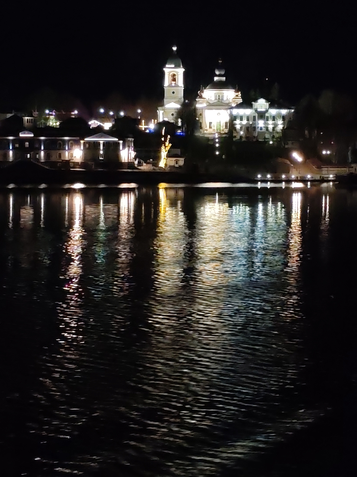 The image shows a nighttime view across a body of water, likely a river, in Nesterovo, Slobodskoe S/P, Russia. Buildings with illuminated exteriors line the opposite bank, including what appears to be a church or cathedral with a prominent bell tower. Reflections of the lights from the buildings are visible in the water, creating a rippled pattern. The overall scene is dark, with the water and sky appearing black, contrasting with the illuminated structures.
