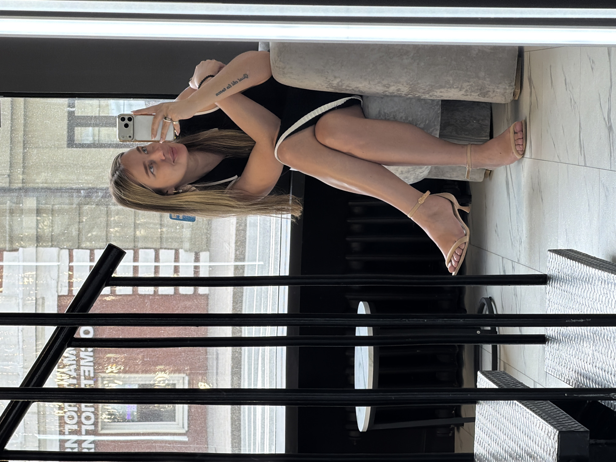 A woman is posing for a selfie in what appears to be an indoor setting with a view of the city of Moscow, Russia. The Moscow Holida Inn can be seen in the background through a window covered with black horizontal bars and what appears to be some form of a metallic or grid-like structure. The woman is seated on a gray-upholstered surface. She is wearing a black outfit with white trim and nude-colored high-heeled sandals. She holds a smartphone up to take a photo of herself. Her right arm is visible, displaying a bracelet and a tattoo. The floor is marble.