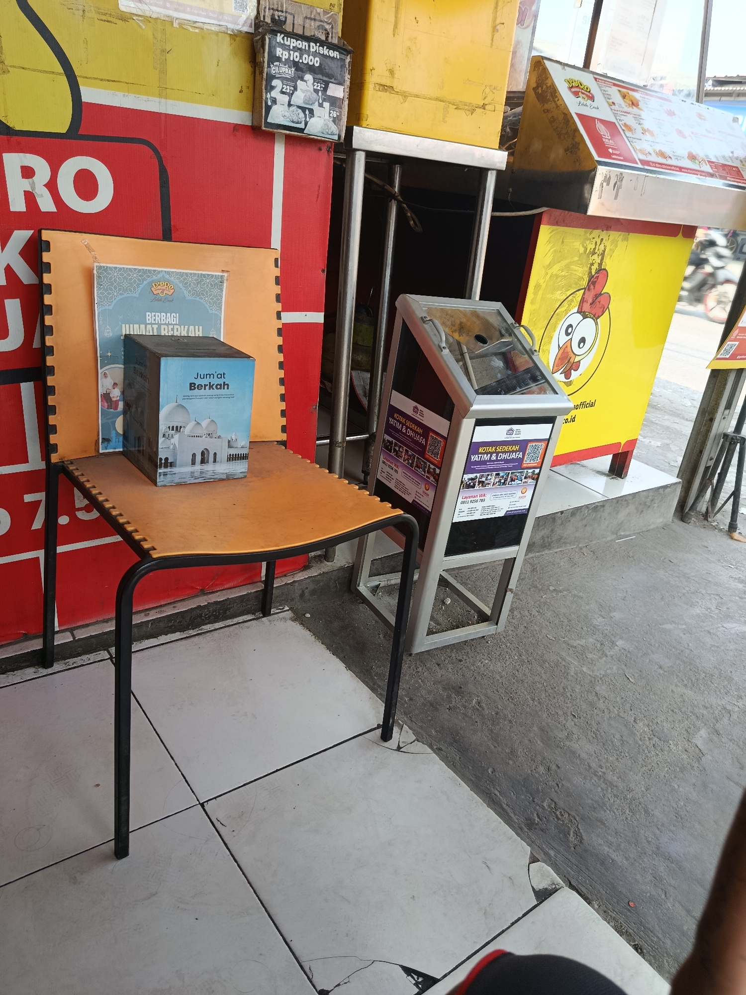 The image shows an outdoor scene, likely in front of a storefront or market stall. In the foreground, a small, orange, kidney-shaped table with black metal legs is positioned at an angle. On the table sits a light blue donation box with an image of a mosque on it, labeled "Jum'at Berkah" (Blessed Friday). Behind the table, a large red sign with white lettering is visible, with the letters "RO" and "K" clearly legible. To the right of the red sign, a yellow sign with a cartoon chicken graphic is displayed. A donation collection kiosk, with a glass top and metallic frame, stands to the right of the yellow sign. The kiosk has posters on its sides advertising donations for orphans and the needy, with QR codes and contact information. In the background, a white tiled floor transitions to a concrete ground. The overall scene suggests a commercial area with promotional materials and a donation drive.