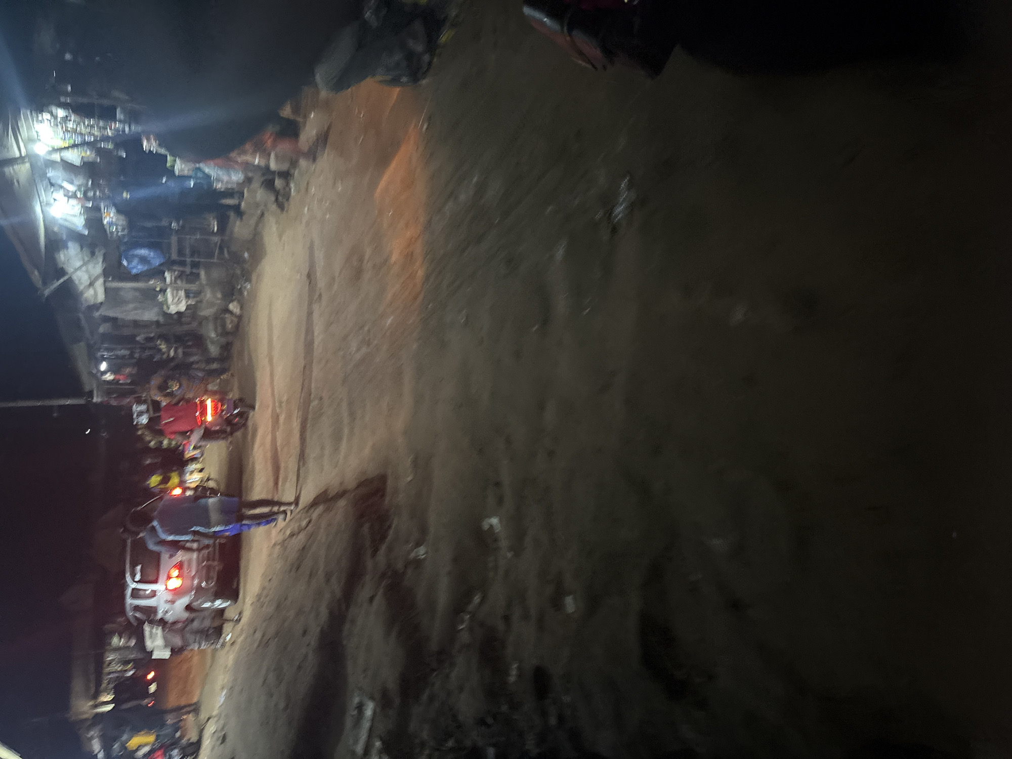 The image depicts a nighttime street scene in Nkpor, Nigeria, characterized by a dirt road lined with makeshift market stalls and a few vehicles. A silver SUV is parked on the left side of the road, its brake lights illuminated. Several individuals are present, with some walking and others on motorcycles. One person in a blue shirt and shorts is walking away from the camera, towards the SUV. Another person in a red shirt is riding a motorcycle with its taillight on, moving away from the viewer. The market stalls on the right are illuminated by artificial lights, revealing various goods displayed for sale. The overall environment appears to be a busy, informal marketplace at night.