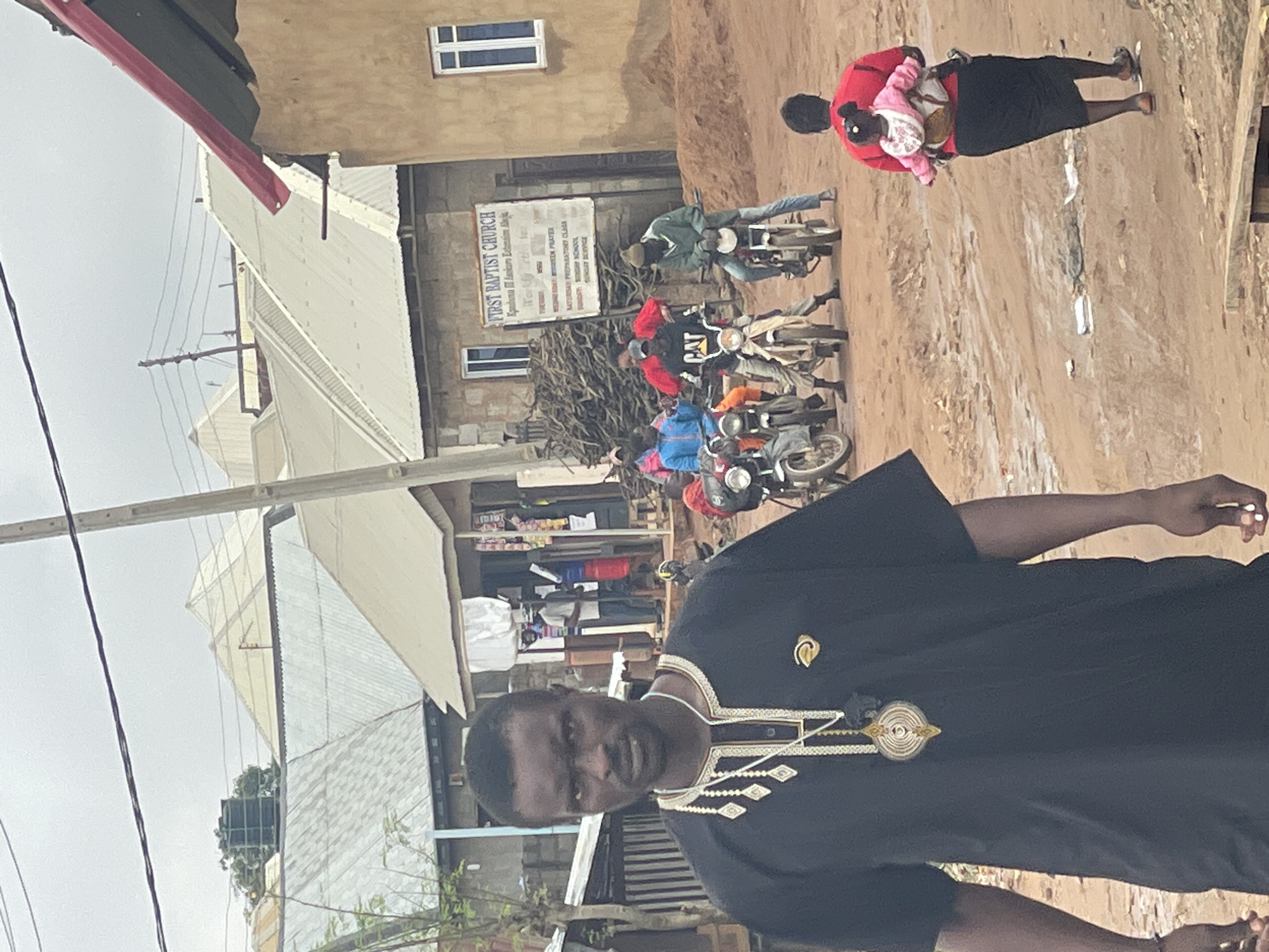 In Kurunduma, Nigeria, a man in a black, embroidered garment stands in the foreground. Behind him, a group of people on motorcycles are gathered near a building with a sign that reads "FIRST BAPTIST CHURCH." The street is unpaved and muddy. In the background, various buildings with corrugated metal roofs are visible. A woman carrying a child on her back walks away from the camera. The sky is overcast.