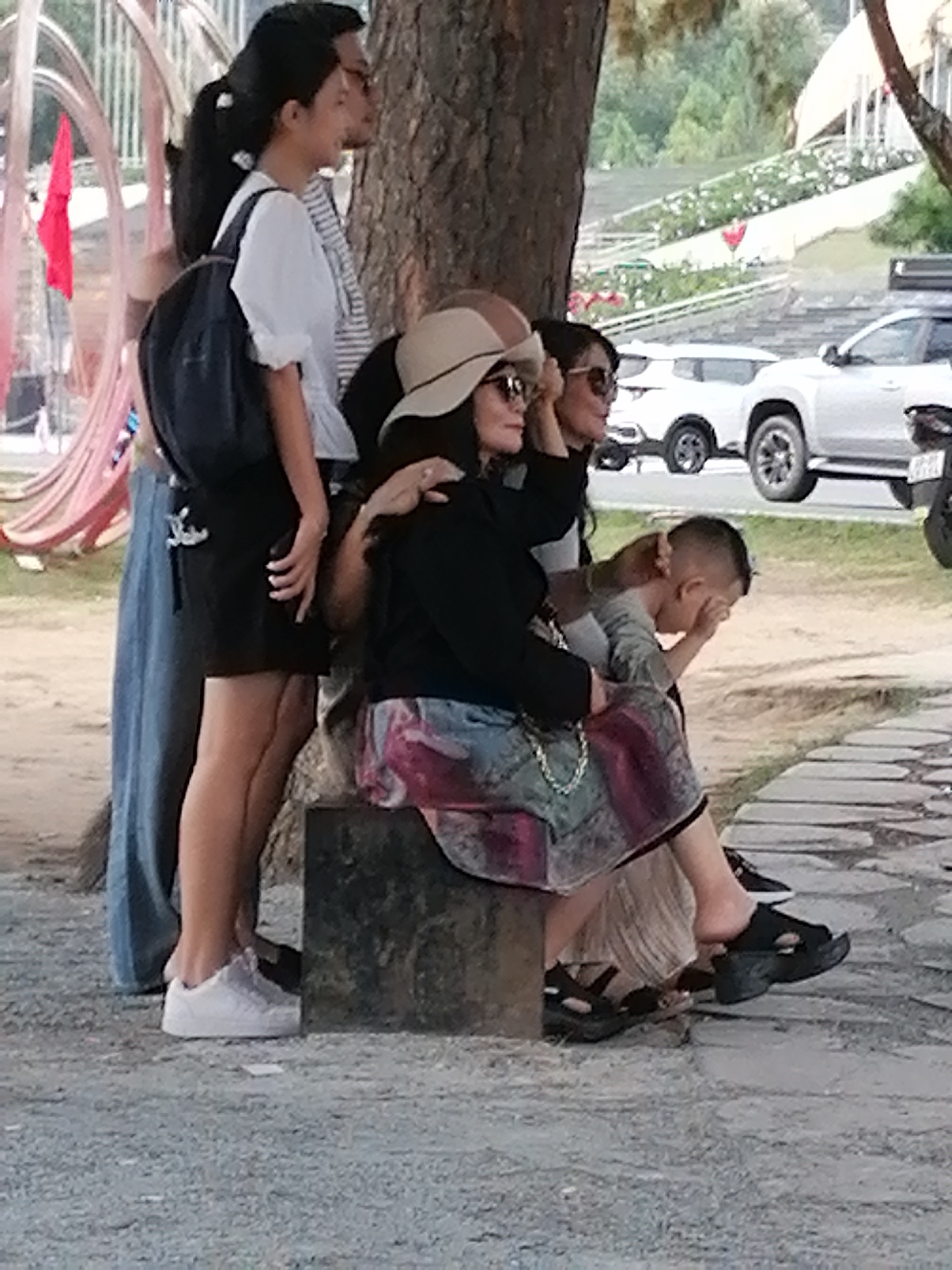 In Da Lat, Vietnam, an outdoor scene unfolds with a group of people near a tree. A woman with dark hair pulled back in a ponytail, wearing a white top and black skirt with a backpack, stands to the left. Next to her, a woman in a tan hat and sunglasses is seated. A child with short hair, wearing a gray shirt, is also seated. In the background, a white SUV and a motorcycle are visible. The ground is covered in gravel, and a paved walkway is present. A red flag is visible on a pink sculpture to the far left.