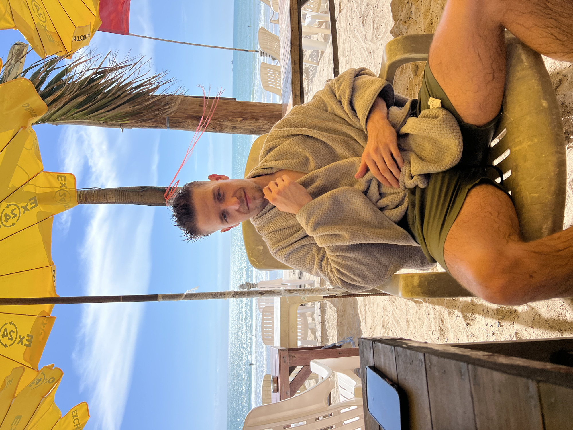 A man sits in a chair on a sandy beach, looking at the camera. He is wearing a gray robe and green shorts. The beach has several tables and chairs, and yellow umbrellas are visible in the background. The ocean is in the distance, with a clear blue sky above. A cell phone rests on a wooden table in the foreground.