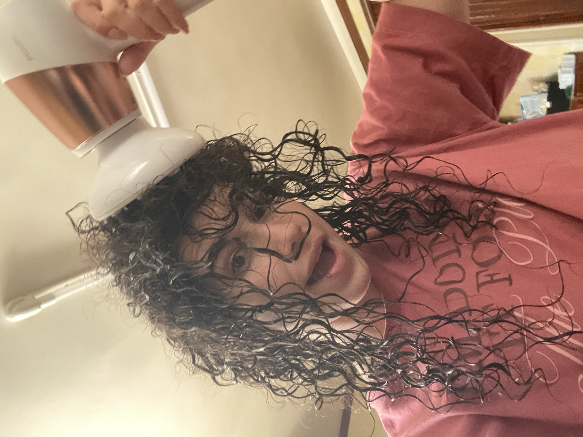 A person with curly, wet hair is drying their hair with a Philips hair dryer with a diffuser attachment. The person is wearing a pink shirt. The photo is taken from a low angle, looking up at the person. The background includes a light-colored wall and a door frame. The location is not discernible from the given image and cannot be identified as Sofia, Bulgaria.