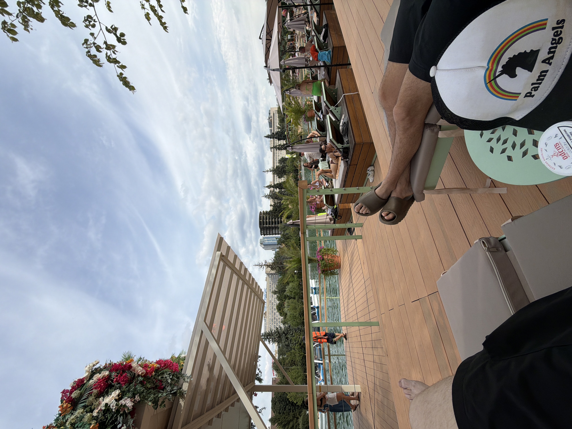 The image depicts an outdoor seating area at a waterfront establishment, likely a cafe or lounge, in Moscow, Russia. In the foreground, a person's legs are visible, wearing black shorts and brown slides, resting on a beige chaise lounge. A black and white baseball cap with a rainbow and unicorn logo, and the text "Palm Angels," rests on a light green side table.
The scene extends to a wooden deck with more chaise lounges, some occupied by individuals sunbathing. A railing separates the deck from a body of water where several small boats, possibly for rent, are moored. A woman in a blue dress and a person in an orange safety vest are visible on the deck near the water's edge. The background shows a line of trees, buildings, and a cloudy sky. The architecture of the buildings suggests an urban environment. The overall atmosphere is relaxed, indicating a recreational setting.