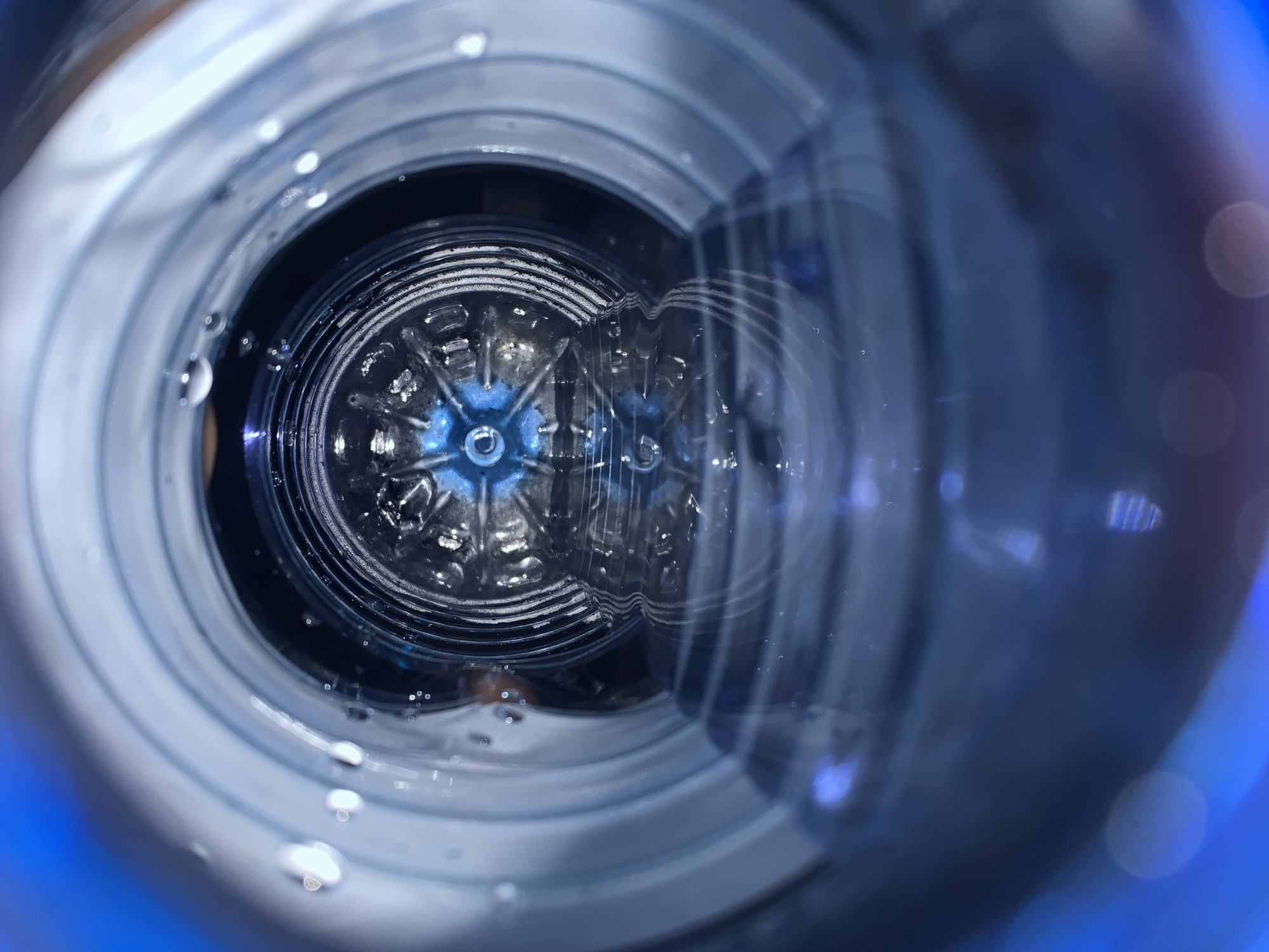 The image shows the interior of a blue plastic water bottle, viewed from the bottle's opening. Concentric rings of plastic form the bottle's structure. At the bottom of the bottle, a star-shaped design is visible. Droplets of condensation adhere to the inner surfaces of the bottle. The image appears to be taken with a close-up perspective. The location of New Stadium, Nigeria is not discernible from the given image.