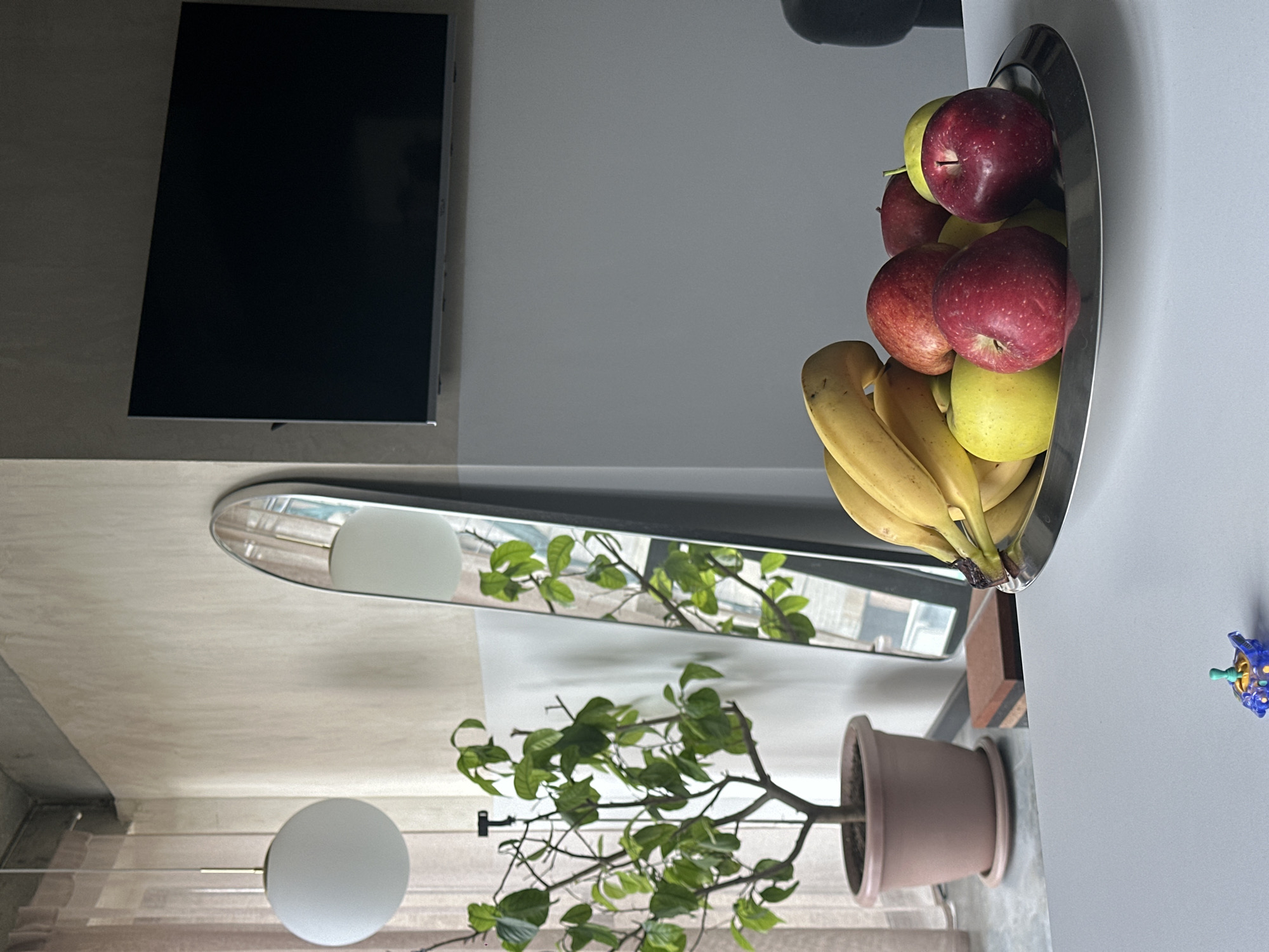 This image captures an interior scene, likely a modern living or dining space in Yerevan, Armenia, characterized by minimalist design and natural elements. The perspective is slightly elevated, looking down towards a surface on the right and across towards a wall on the left.
On the right side of the frame, a polished silver fruit bowl rests on a light grey table or counter. The bowl is generously filled with a variety of fresh fruit, including several vibrant red apples, some green or yellow apples, and a bunch of ripe yellow bananas. A small, intricate decorative object, appearing blue and green with a spiky texture, is situated on the same surface to the right of the fruit bowl.
To the left, a large, black flat-screen television is mounted on a light beige or off-white wall. Below the television, a tall, narrow mirror with rounded ends is mounted vertically on the same wall. The mirror reflects parts of the room, including green foliage from a plant and a spherical white light fixture, suggesting a window or source of natural light might be opposite it. Below the mirror, a potted plant with delicate green leaves and thin branches stands in a light brown terracotta pot.
The overall lighting appears soft and natural, indicative of daytime, though specific weather conditions are not discernible. The scene is static, devoid of any human presence or discernible activity. No visible text is present in the image.