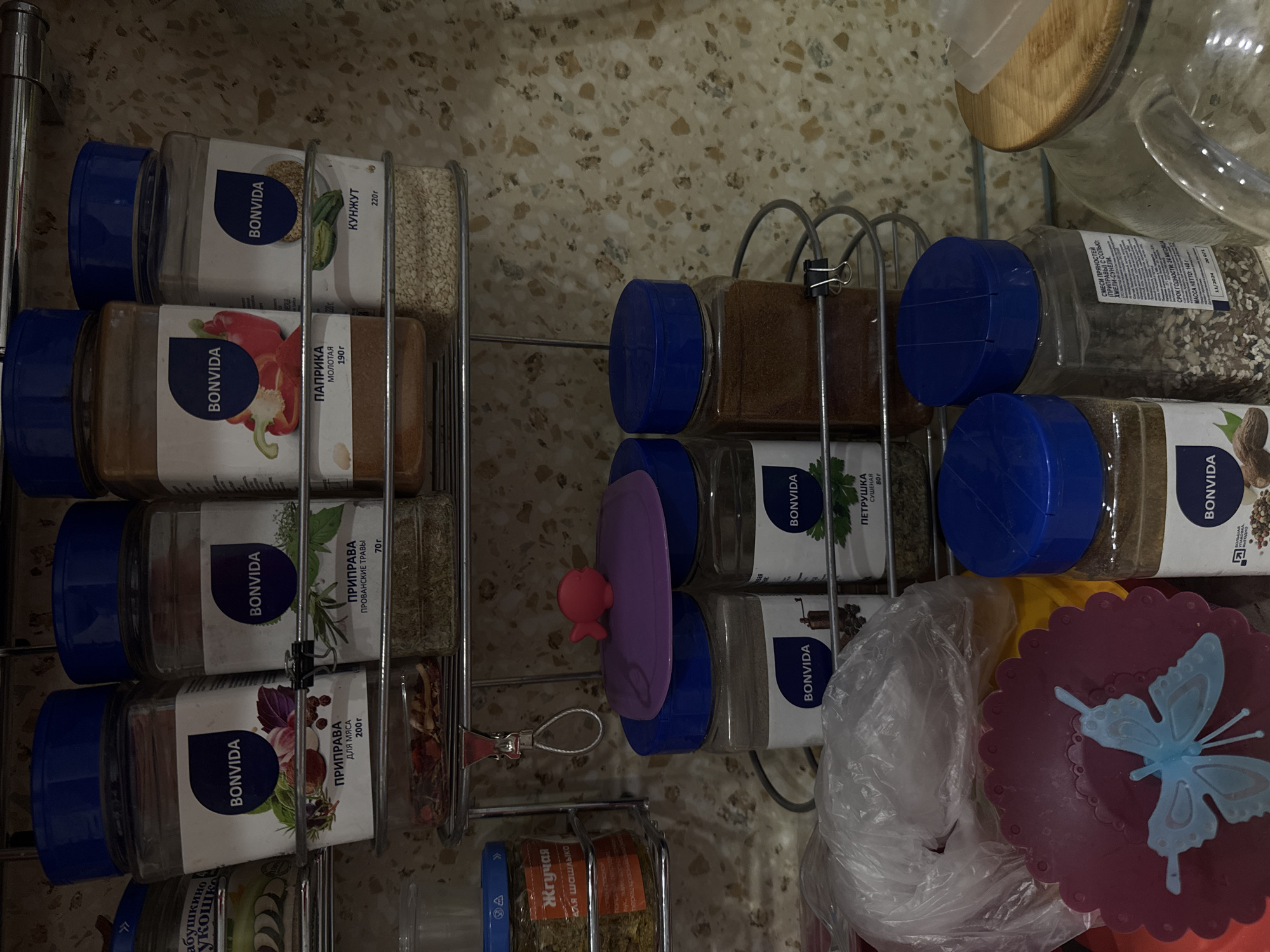 The image shows a collection of spice jars arranged in a metal spice rack, likely in a kitchen. The jars are mostly labeled "BONVIDA" with various spices, some labeled in Russian, such as "Кунжут" (sesame), "Паприка" (paprika), "Петрушка" (parsley), "Прованские травы" (Provencal herbs), and spices for meat or shashlik. A pink silicone lid and a white plastic bag are also visible, and a glass jar with a wooden lid. The label on one jar indicates the spices are a "смеси пряностей" (spice mix), specifically "Хмели-сунели" (khmeli suneli) with an expiration date of 11/2024 and a net weight of 140 grams. While the spice labels and a visible "абушкино лукошко" ingredient, point to the contents, a precise geolocation within Izhevsk, Russia, is not possible from the image.