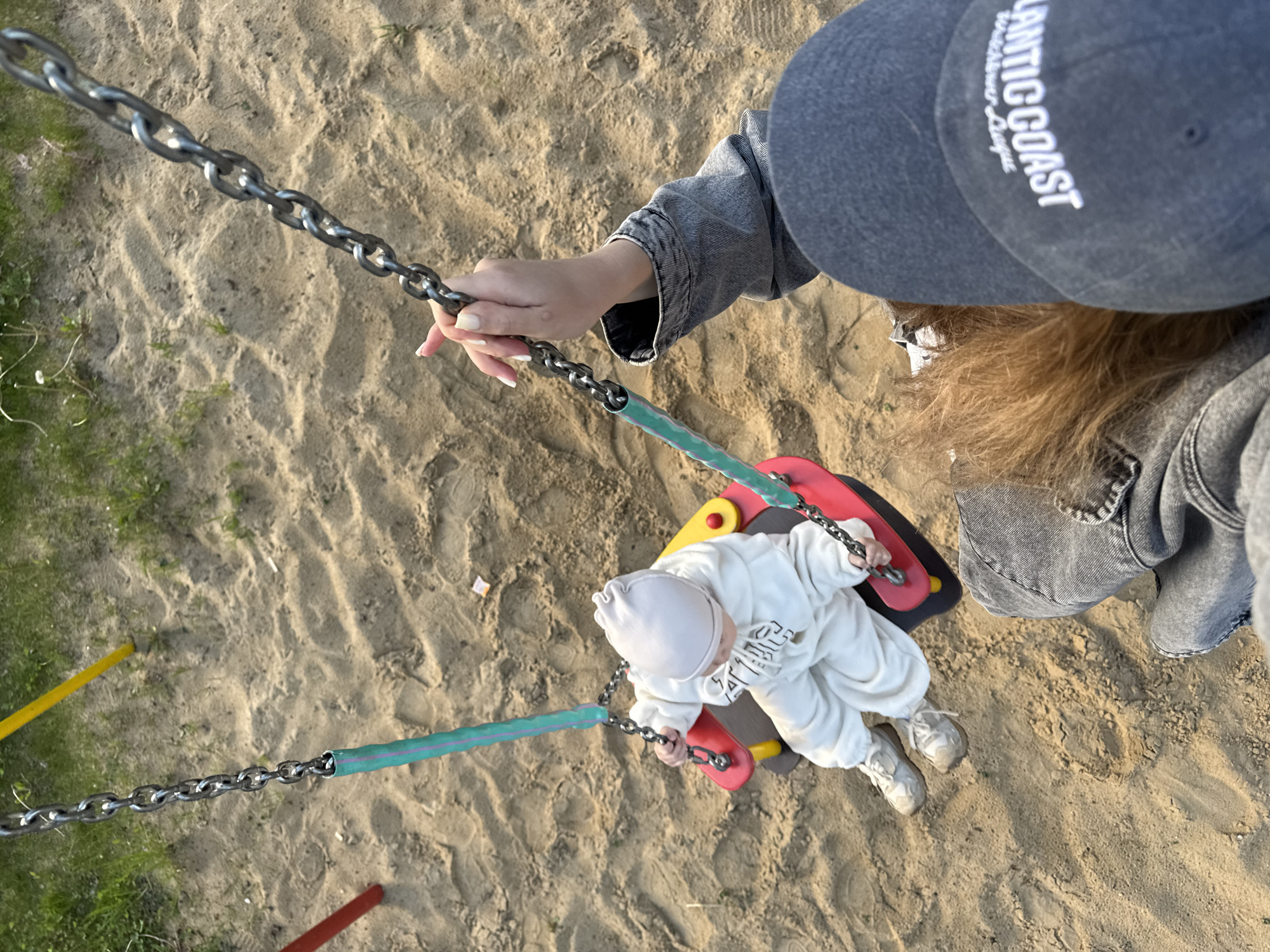 A top-down shot shows a child on a swing set in a sandy area, likely a playground. The child, wearing a white outfit and matching hat and white sneakers, sits in the swing seat. A person with a gray cap and gray denim jacket is holding the swing chain with their left hand. The location may be Vladimir, Russia based on the user request.