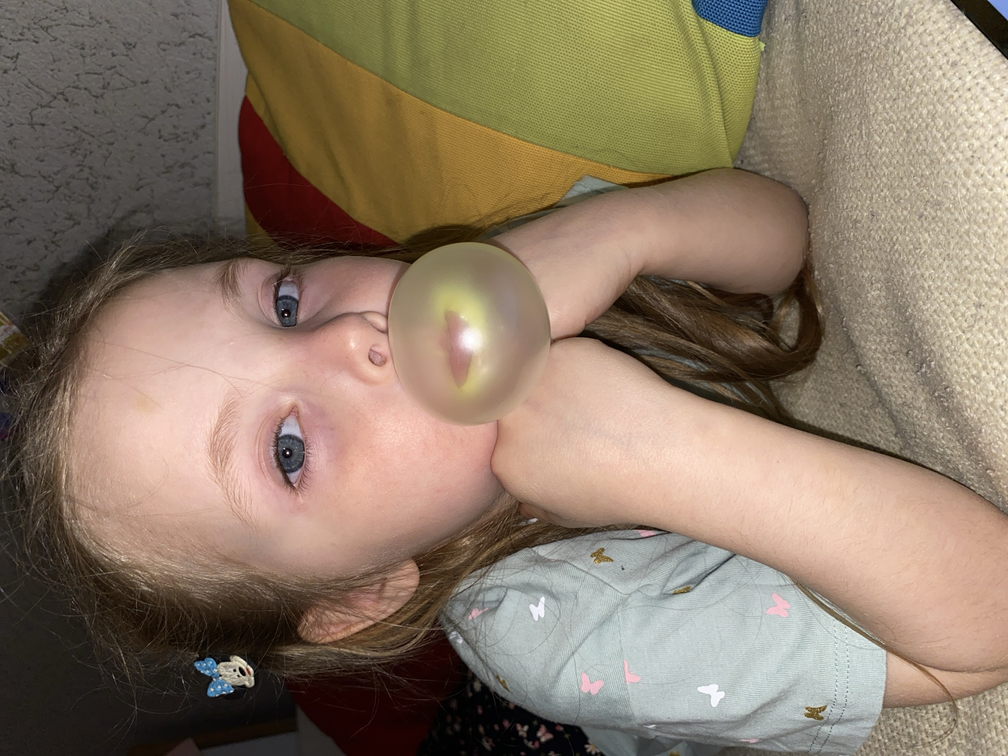 This close-up, angled portrait captures a young girl, possibly in Votkinsk, Russia, lying down and blowing a large, translucent chewing gum bubble. The bubble, which has a yellowish-pink sheen, is positioned centrally in front of her mouth and nose.
The girl has fair skin, light brown hair, and blue eyes, which are open and looking slightly upward with a focused or contemplative expression. She is wearing a light blue-green t-shirt decorated with small pink and gold butterfly patterns. A decorative hair clip, shaped like a blue and white bow with a white character's head, is visible in her hair near her left ear. Her arms are bent, with her hands positioned near her face.
She is resting against a textured, light beige upholstered surface, likely a couch or cushion. Behind her head and upper body, a vibrant, striped fabric item—possibly a blanket or pillow—displays sections of green, yellow, red, and blue. The indoor lighting suggests an artificial light source. No visible text is present in the image.