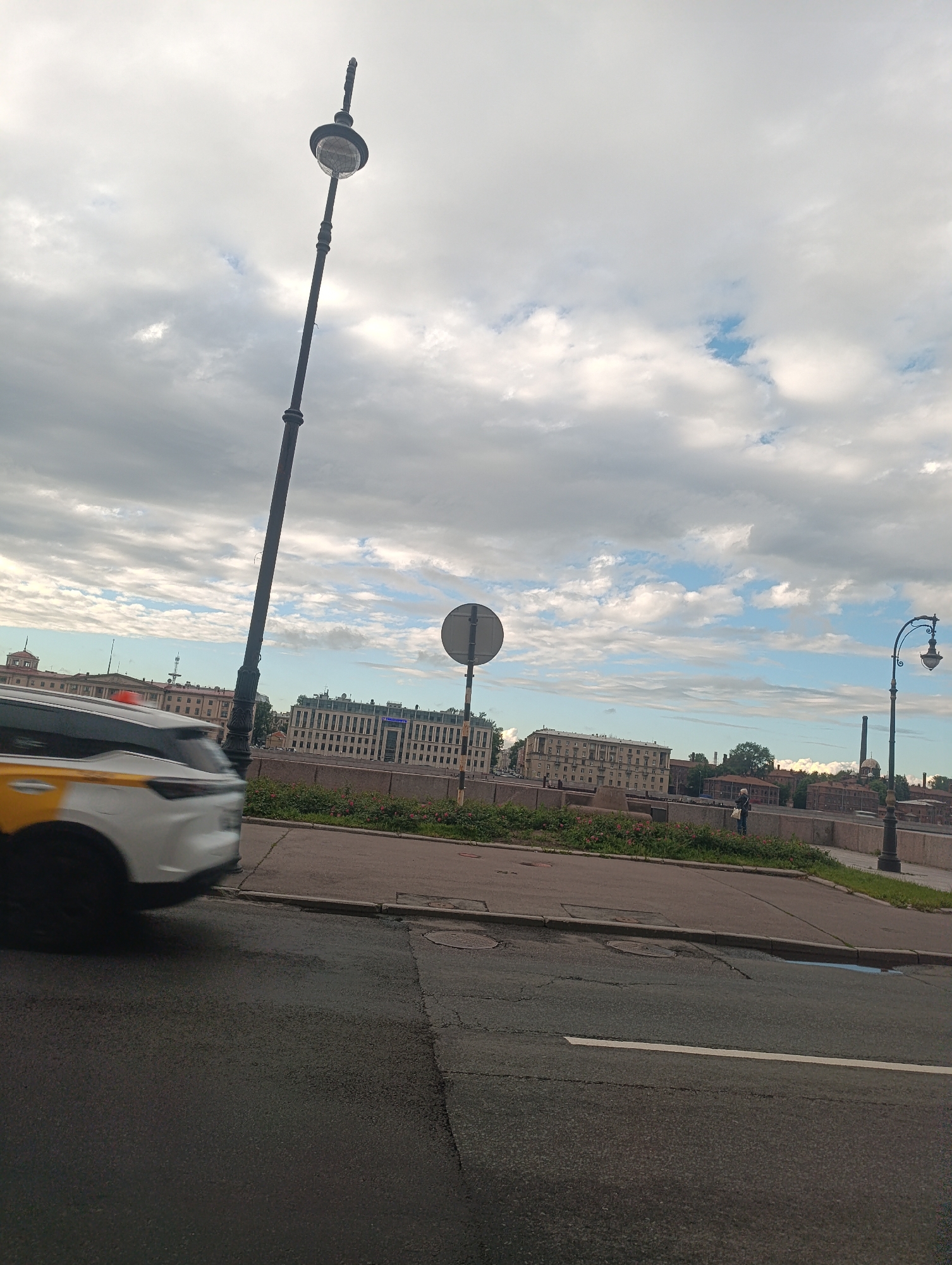 This image depicts a cloudy day in Saint Petersburg, Russia, likely in the late afternoon or early evening due to the soft light. The scene is a street view, with a focus on a tall, ornate lamppost dominating the left side of the frame. In the background, a row of multi-story buildings lines the riverbank, suggesting an urban setting. A white and yellow car, possibly a taxi, is captured in motion, blurred due to its speed, moving from left to right in the foreground.
Further to the right, another lamppost with a more curved design is visible, adding to the architectural character of the street. A single person stands on the riverbank, appearing to be observing the surroundings. The sky is filled with dramatic clouds, interspersed with patches of blue, indicating a dynamic weather pattern. The road in the foreground shows signs of wear, with cracks and manhole covers. The overall atmosphere is one of urban life unfolding under a changeable sky. No visible text is present in the image.