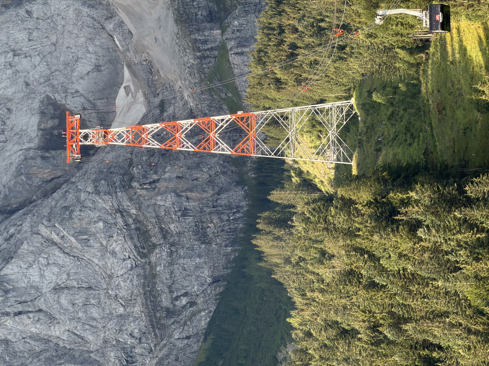 An aerial tramway tower, painted red and white, stands in a mountainous landscape. The tower is made of metal lattice work and is topped with a red platform. It is surrounded by lush green pine trees that cover the slopes of the mountain. In the background, a massive gray rock face rises steeply. A single aerial tramway car, black with a red logo, is visible suspended from cables, moving towards the right of the frame. The scene is bathed in natural sunlight, creating shadows and highlighting the textures of the rocks and trees. The location is the Zugspitze cable car station in Grainau, Germany, as indicated by the logo on the tram car.