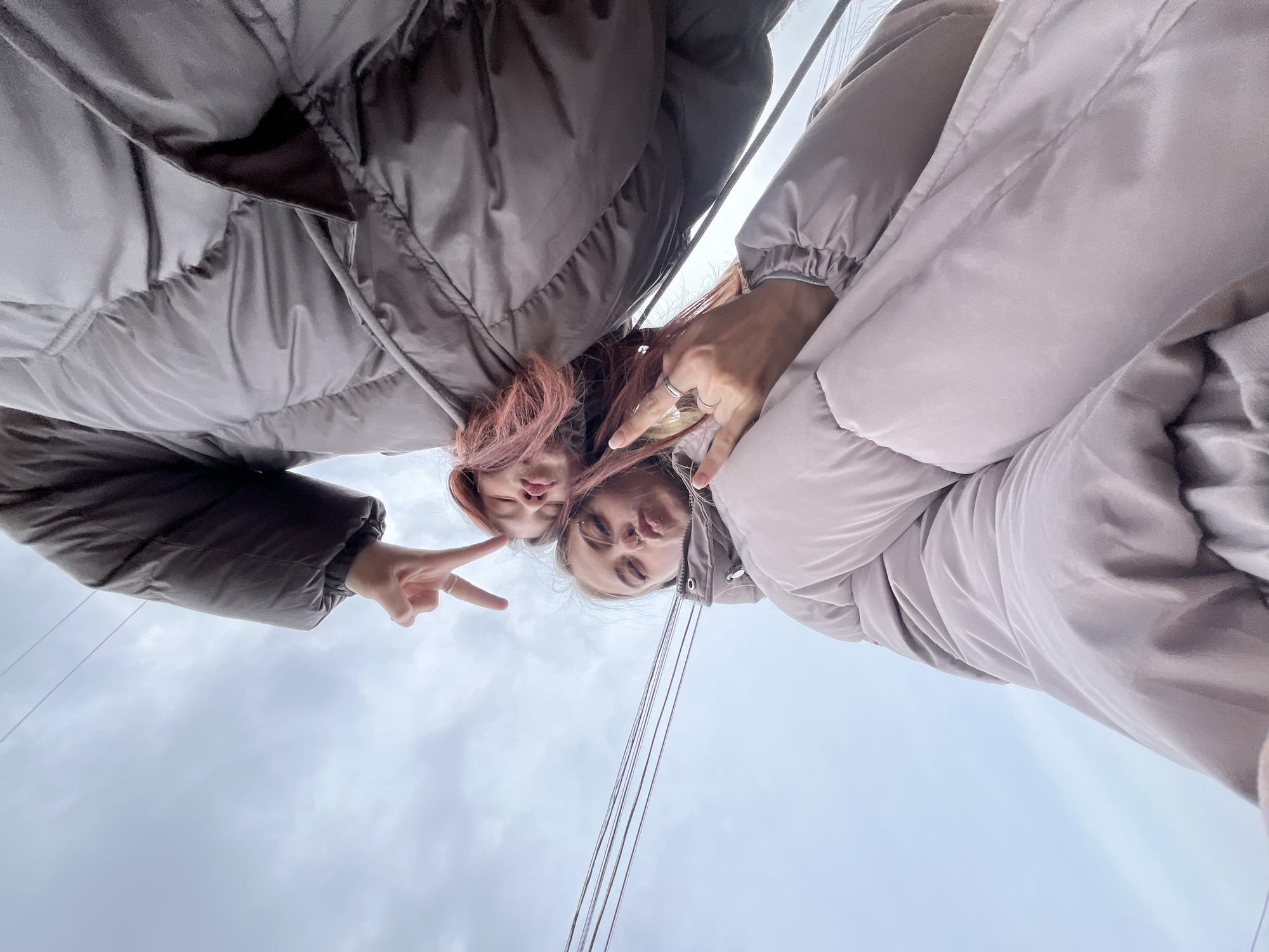 Two individuals are positioned closely together, facing downward towards the camera, appearing to take a selfie. The person on the left has pink hair and is making a peace sign with their hand. The person on the right is making a duck face. Both individuals are wearing puffy jackets. Overhead electrical wires run across the frame. The sky is overcast. The location is Фоки, Russia.
