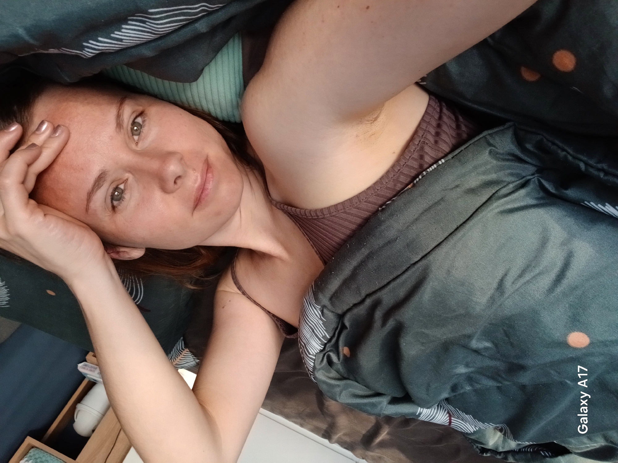 A woman is lying in bed, partially covered by a dark green patterned duvet. She is facing the camera with her head propped up by her hand. She is wearing a brown ribbed tank top. Her arm is raised, revealing a hairy armpit. A bedside table with various items is visible in the lower left corner of the image. The location is a private residence, not specifically identifiable as Yekaterinburg, Russia without additional context.