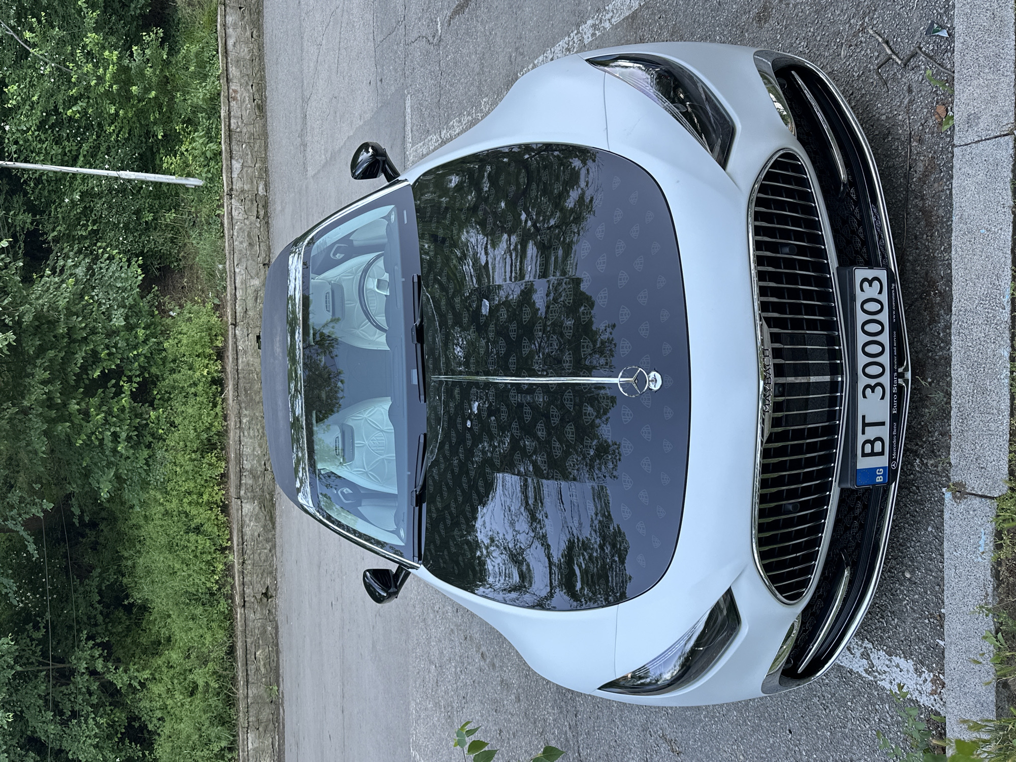 A white Maybach sedan with a black panoramic roof is parked on a concrete surface near greenery, possibly in Veliko Tarnovo, Bulgaria. The vehicle's Bulgarian license plate reads "BT 300003." The roof and interior upholstery of the car feature a repeating pattern of the Maybach logo. The grill of the car faces the viewer. The hood of the car has reflections of trees and sky. The image appears to be taken from an elevated position, looking down on the car.
