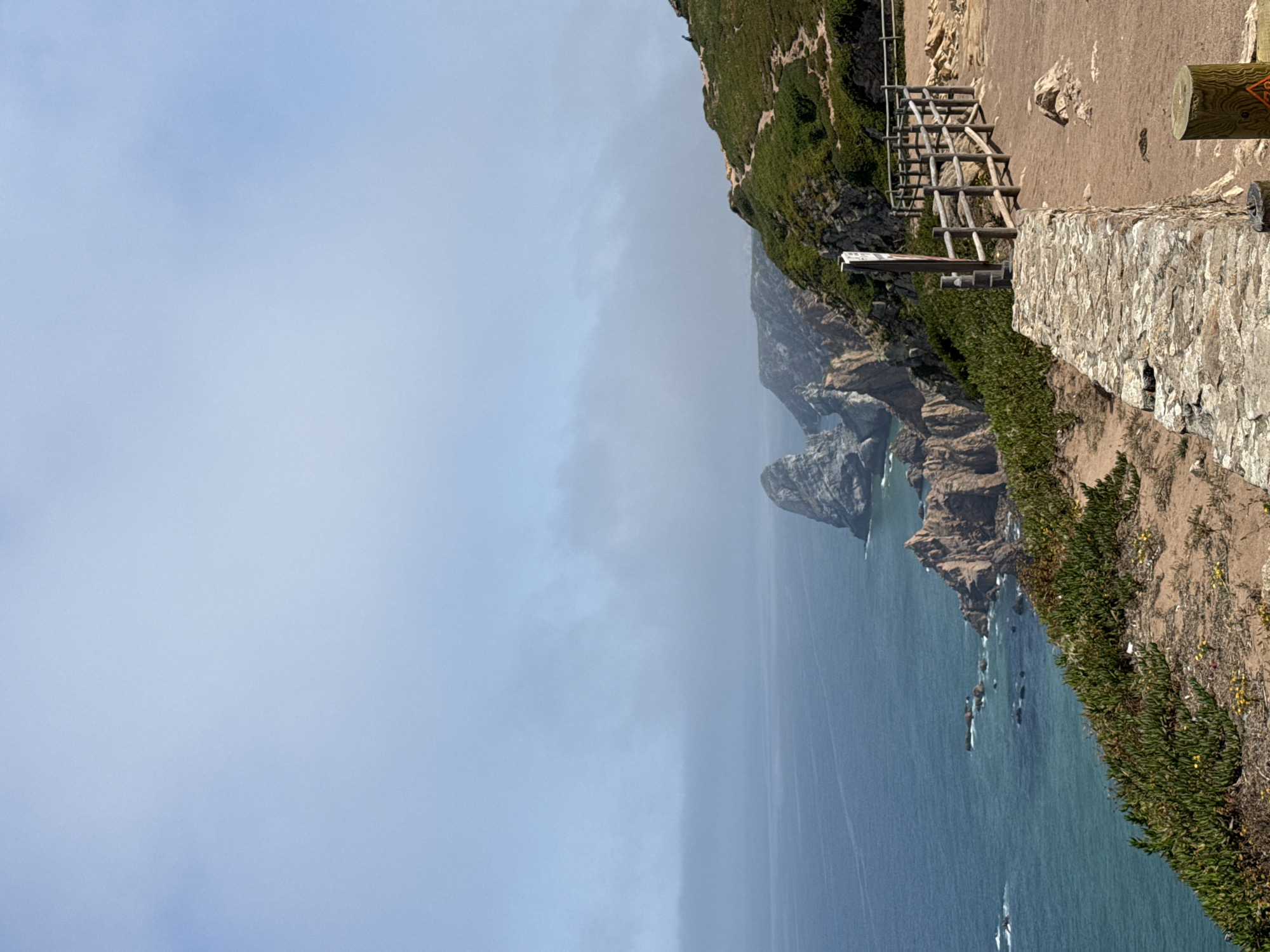 This is a landscape photograph taken at Cabo da Roca, the westernmost point of mainland Europe, near Colares, Portugal. The image captures a dramatic coastal view with rugged cliffs meeting a vast expanse of deep blue ocean. In the foreground, a sandy path runs alongside a rustic wooden fence and a low stone wall. Vegetation, including green succulents and small yellow flowers, grows along the cliff edges. In the distance, several rocky outcrops emerge from the sea. The sky is partly obscured by a light haze or fog, creating a soft, diffused light. No people are clearly visible in the frame, although a distant figure can be seen on the hillside on the right. The overall scene conveys a sense of natural beauty and the raw power of the coastline.